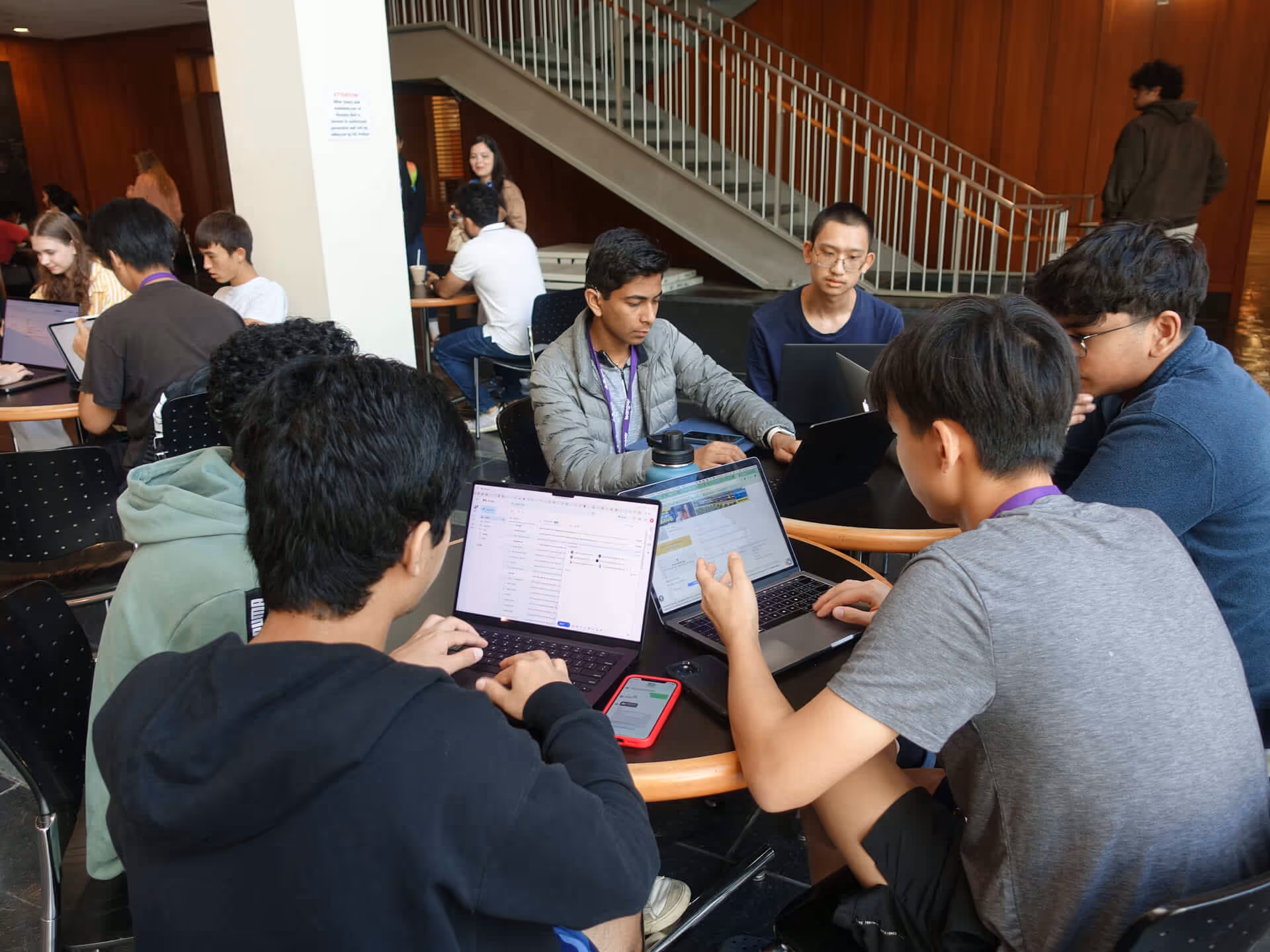 Group of diverse students working on laptops in a study area with a staircase in the background.