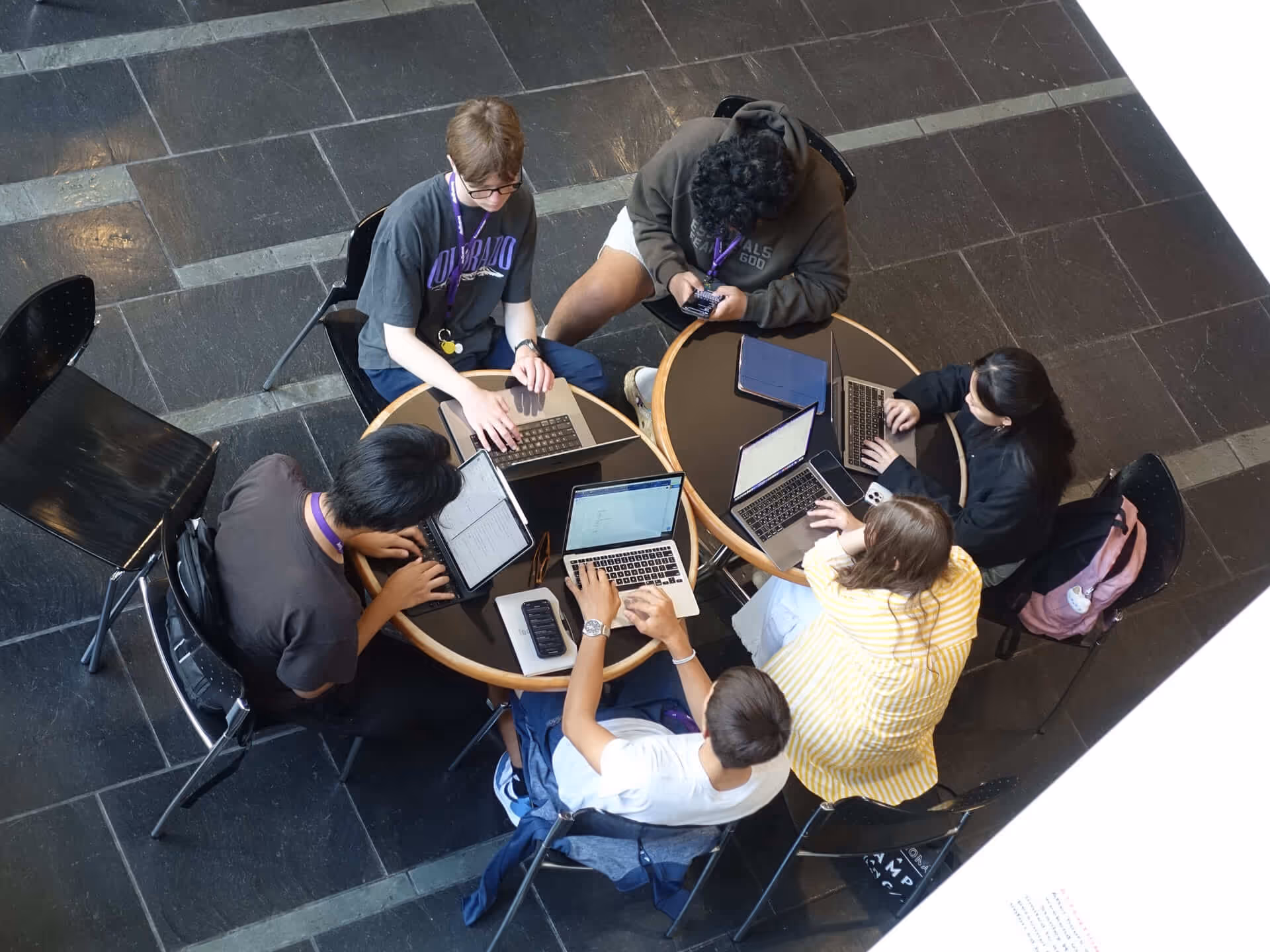 Six teammates sitting around two tables working on laptops and tablets in a tiled floor room.