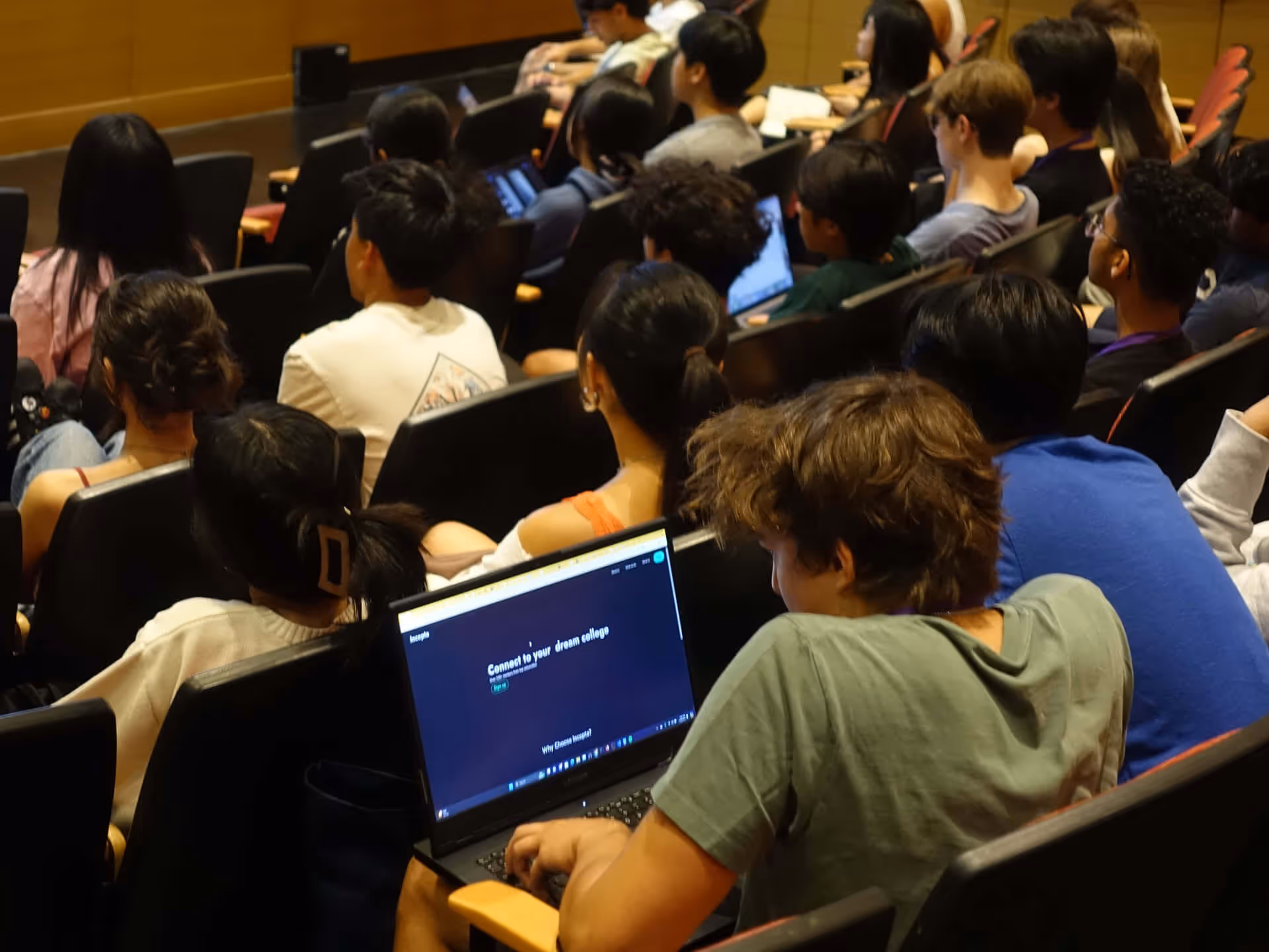 Students seated in a lecture hall, with one focused on a laptop showing a college connection website.