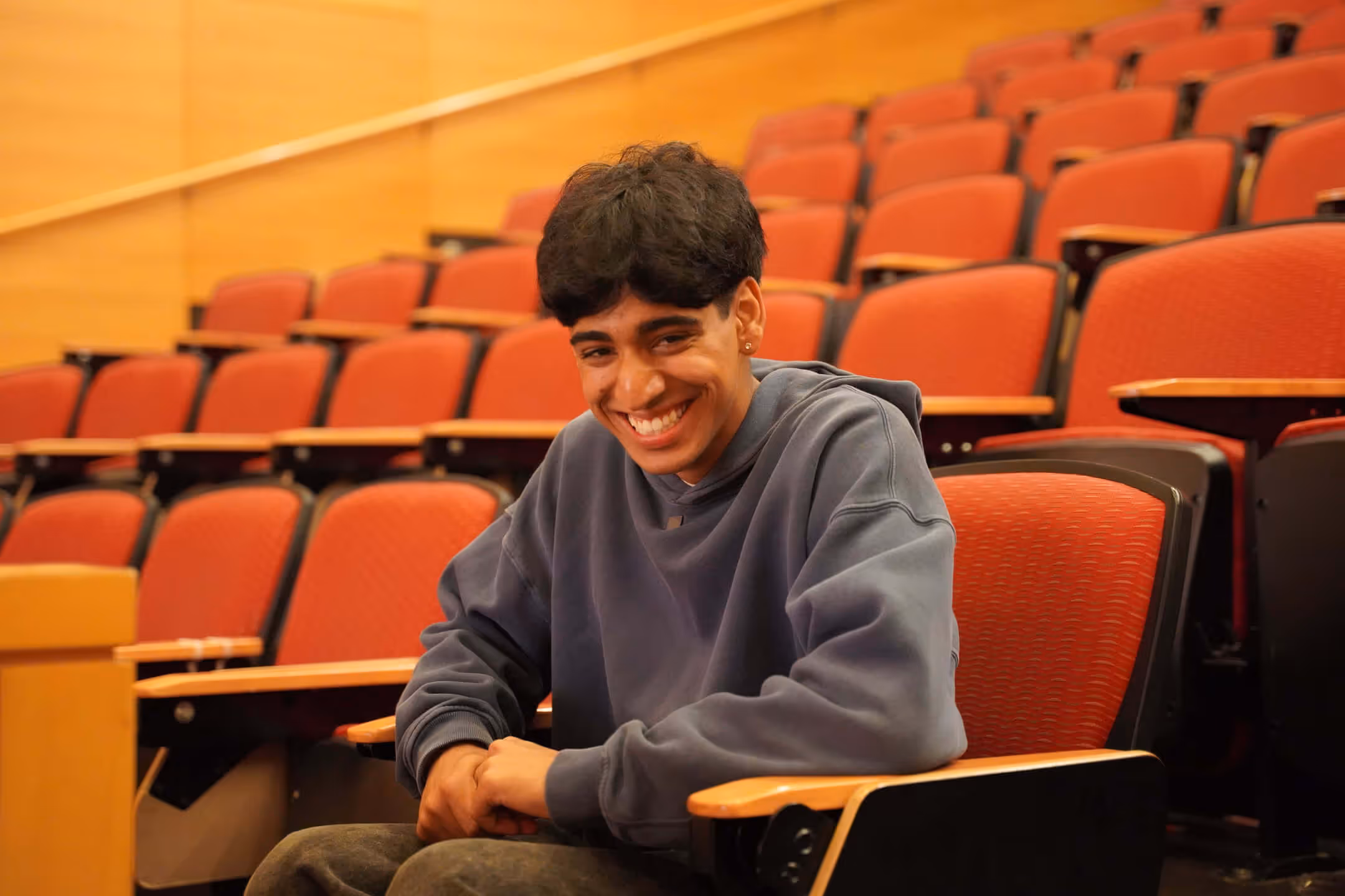 Smiling young man in a gray sweatshirt sitting in an empty lecture hall with red seats.