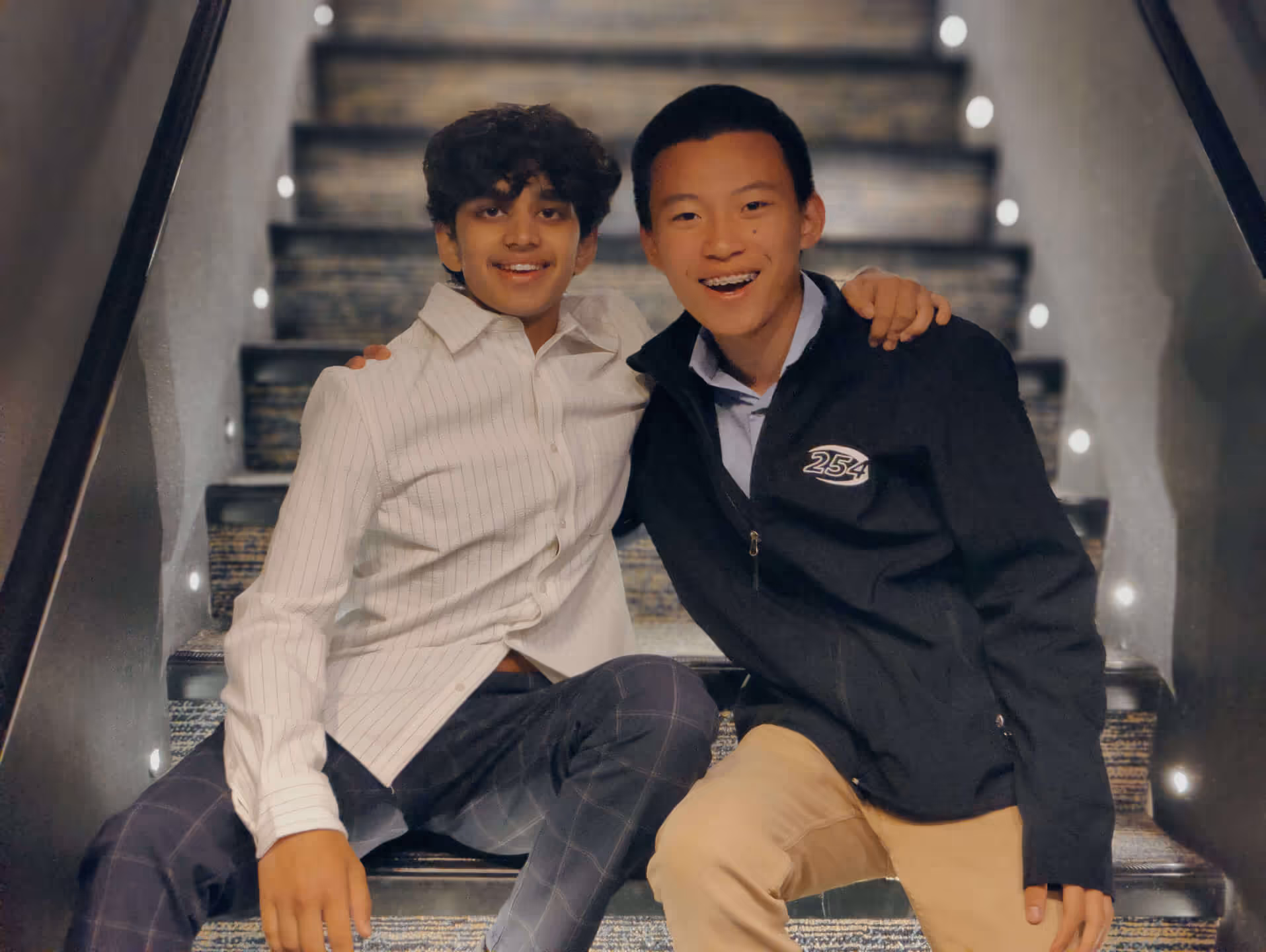 Two teenage boys smiling and sitting on carpeted stairs with string lights along the railing.