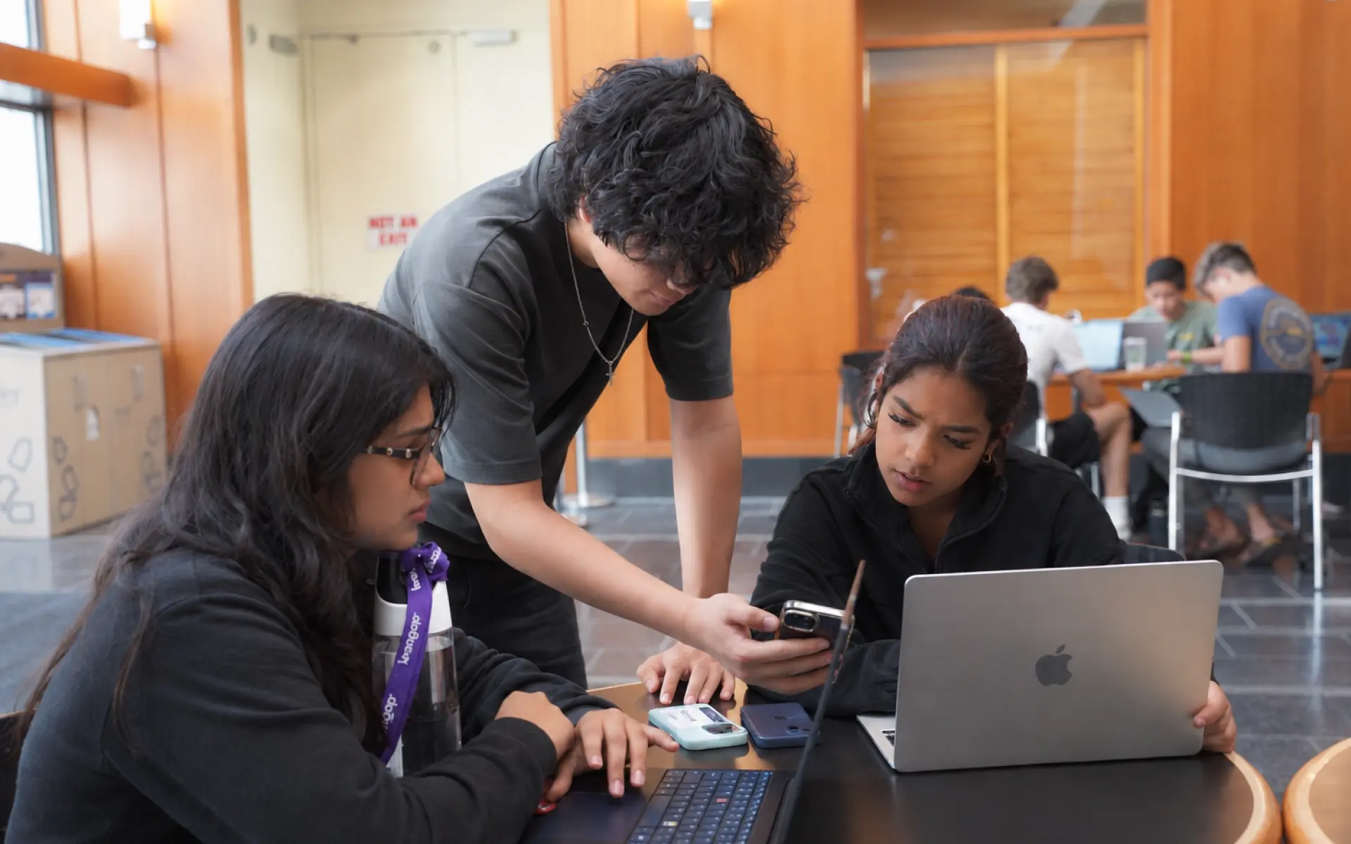 Three teenage students huddled around laptops and phones, testing their startup prototype