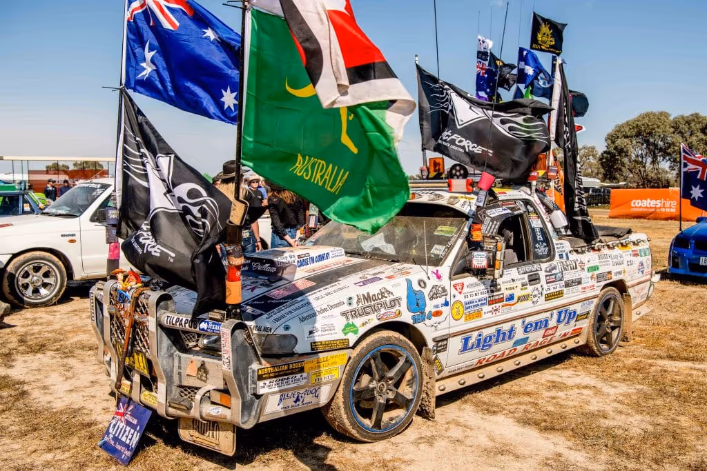 White car decorated with numerous stickers and multiple flags including Australia and a green flag, parked on dry grass at an outdoor event.