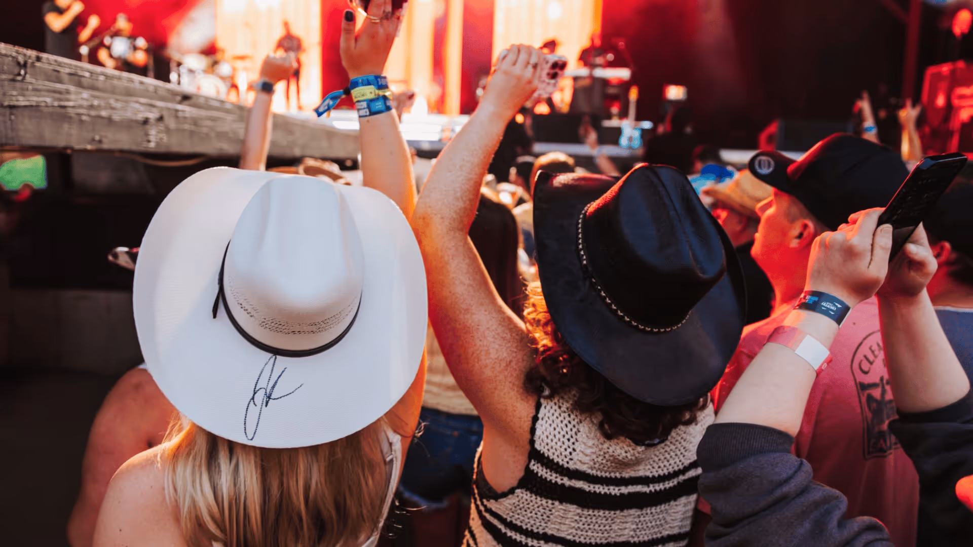 Crowd at an outdoor concert with people wearing hats and raising their hands toward the stage.