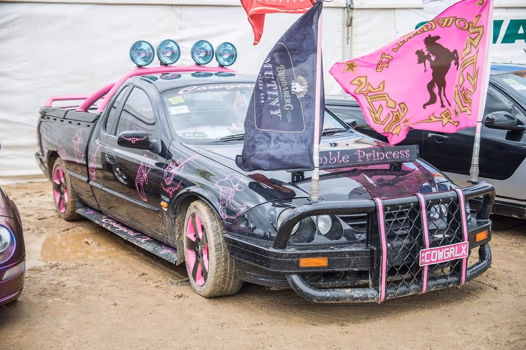 Black pickup truck with pink accents, including wheels, roof lights, and decorations, featuring flags and the name 'Cowgirlx' on the front license plate.