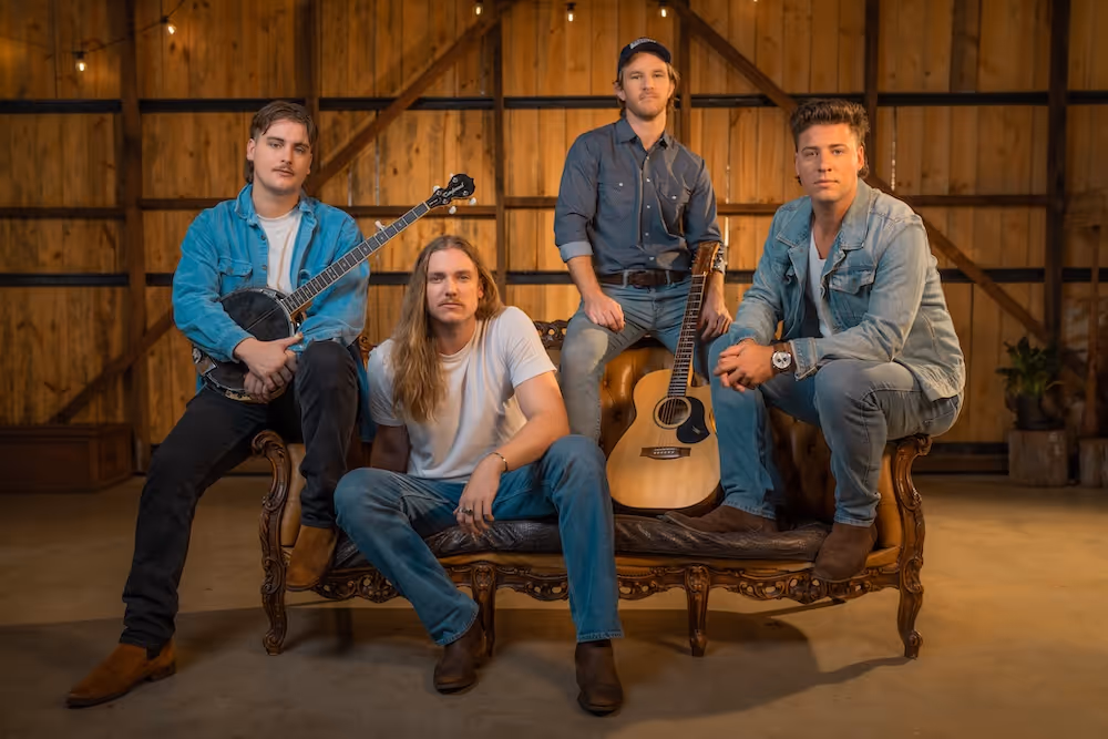The Wet Whistles - four men sitting and standing on a vintage wooden couch in a rustic room, two holding guitars.