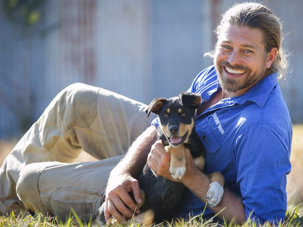 Smiling man in blue shirt lying on grass holding a happy black and tan puppy.