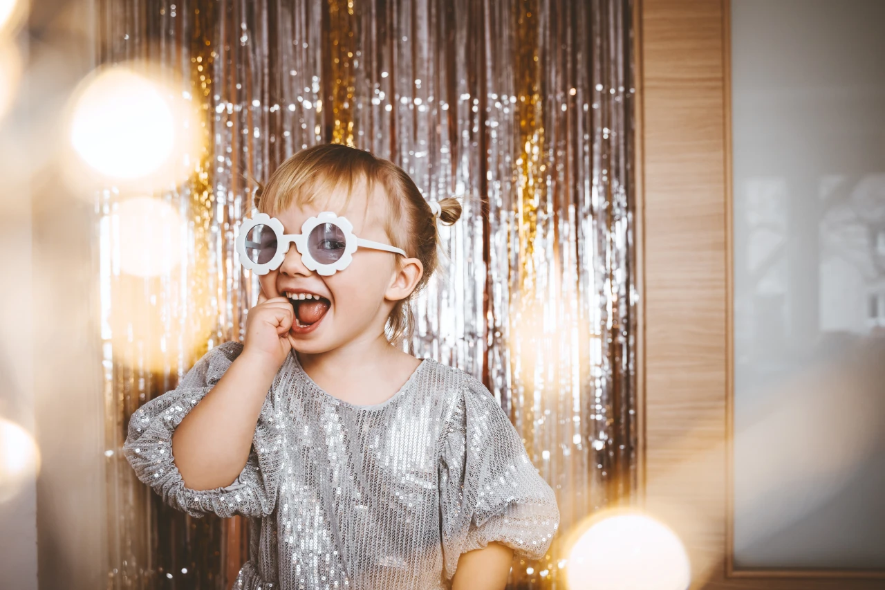 Smiling toddler wearing white flower-shaped sunglasses and a sparkly silver dress at a party backdrop.