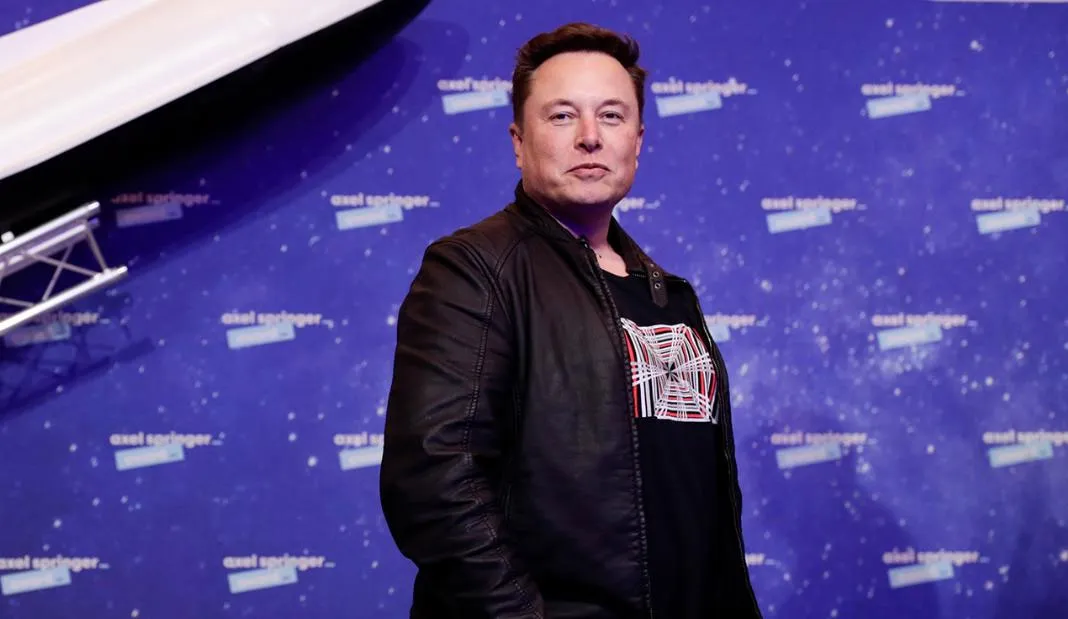 Elon Musk standing confidently, wearing black in front of a blue background.