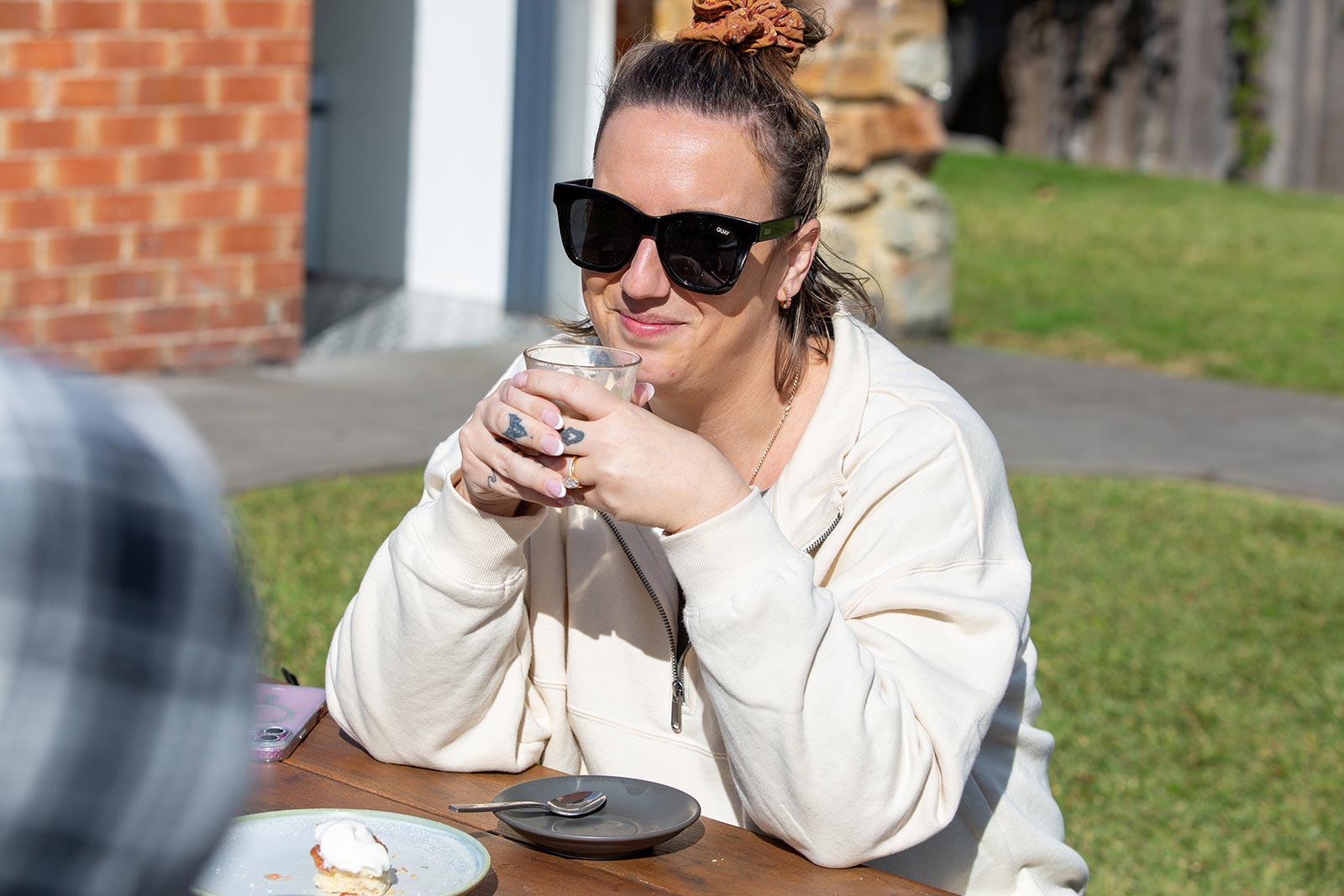 Woman wearing large black sunglasses and a cream hoodie, holding a glass and smiling while sitting outdoors at a wooden table with dessert plates.