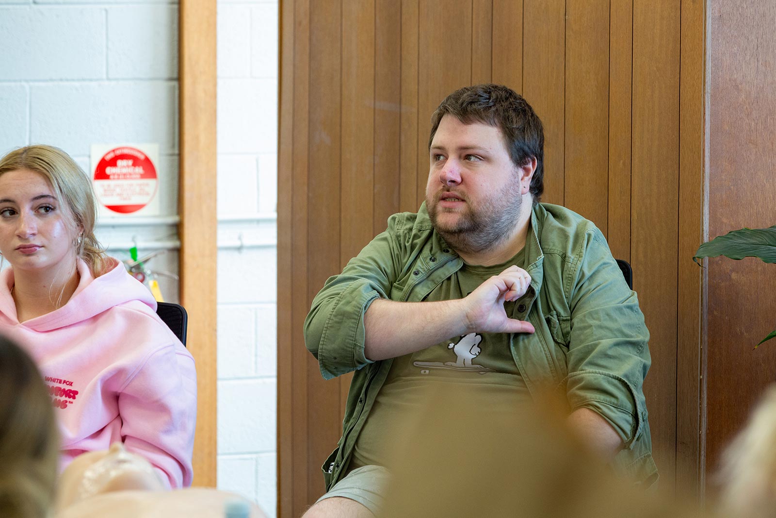 A man in a green shirt and jacket sits next to a woman in a pink hoodie in a room with wooden paneling and a white brick wall.