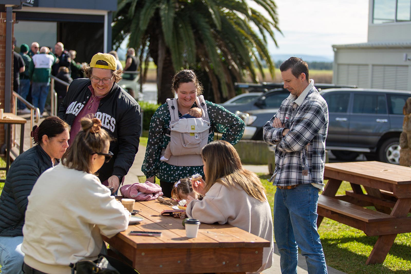 Group of people gathered outdoors around a wooden picnic table, including a woman carrying a baby in a front carrier, with cars and palm trees in the background.