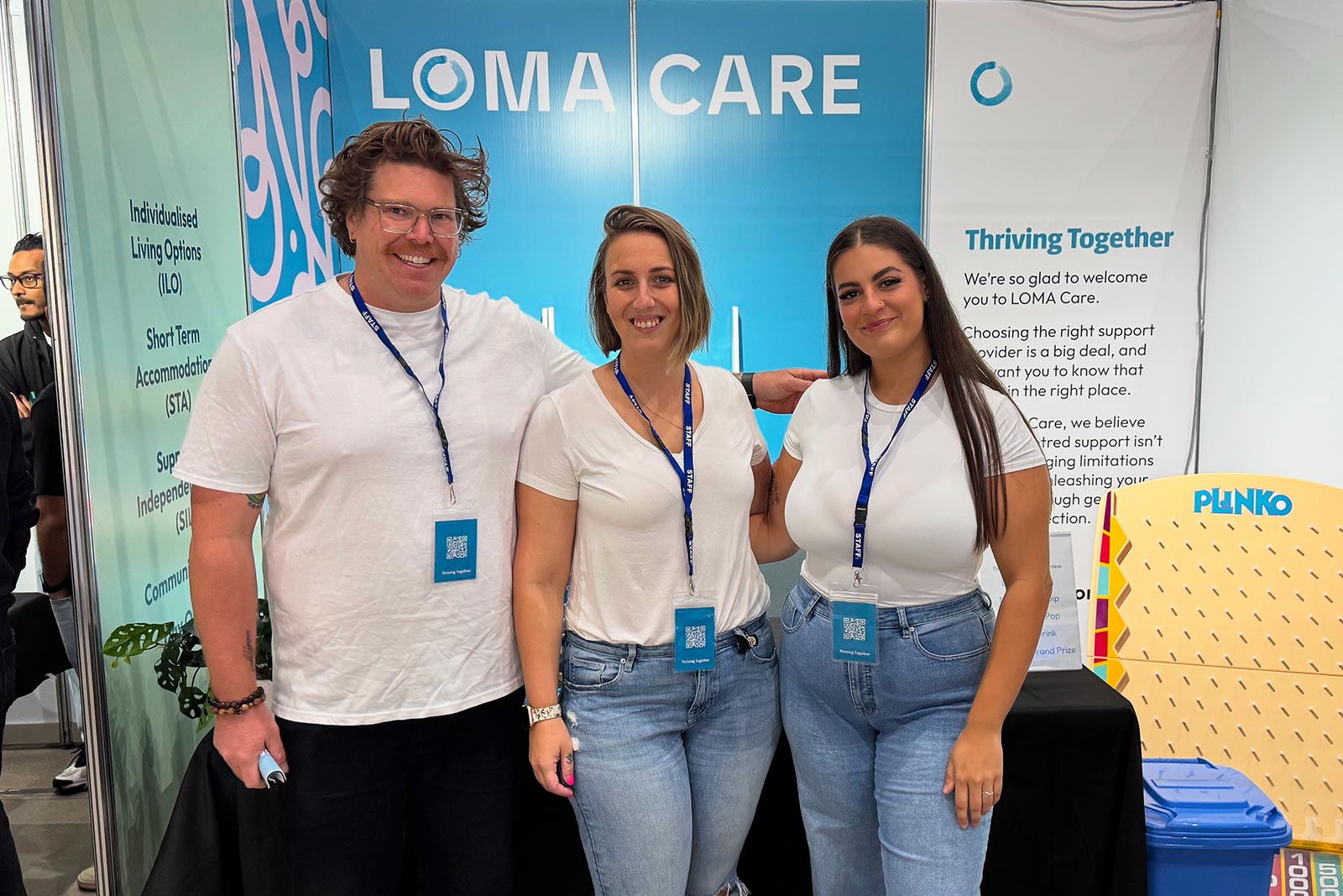 Three smiling LOMA Care staff members wearing white shirts and blue staff badges standing together in front of a LOMA Care display booth.