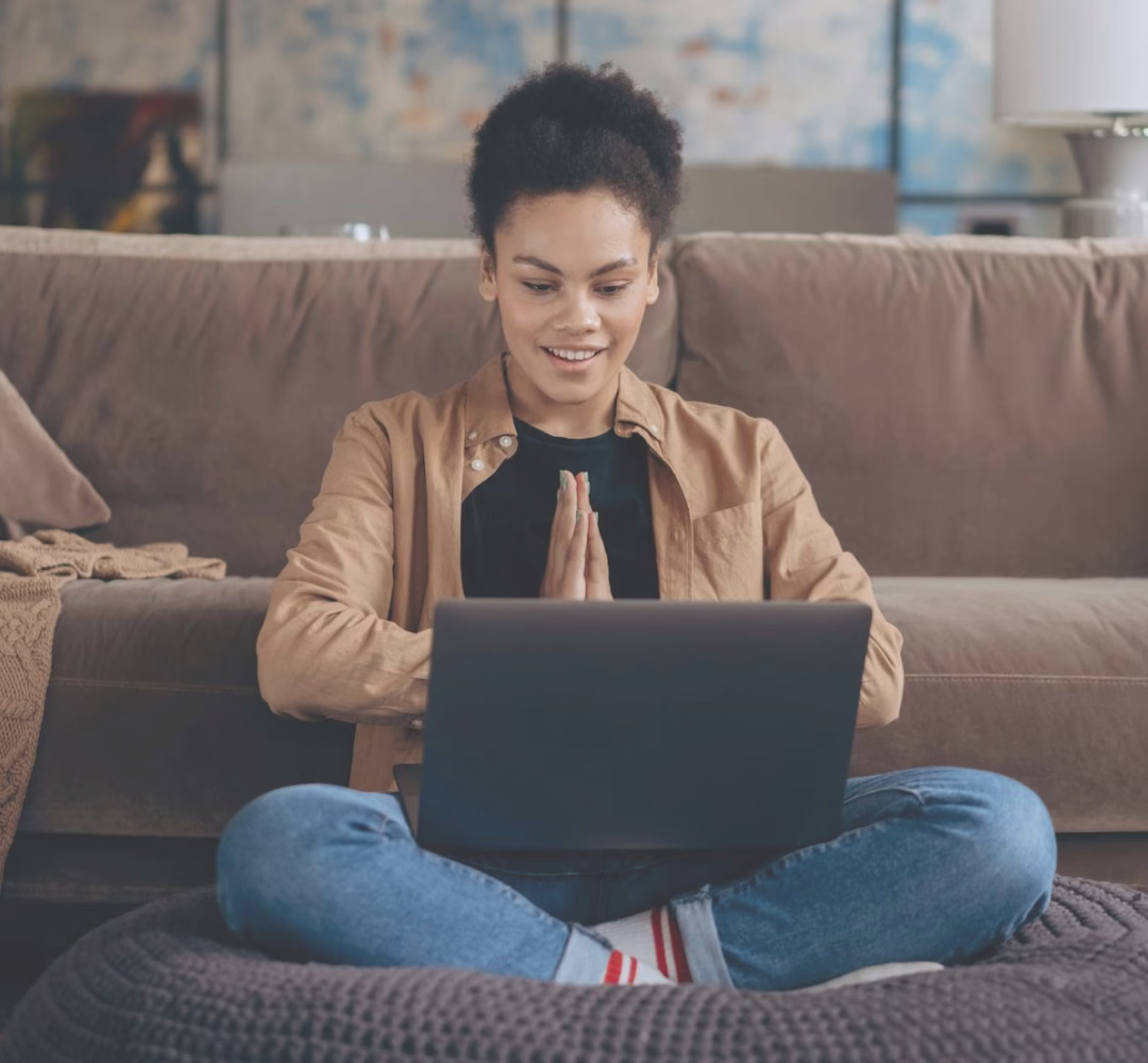 Young woman sitting cross-legged on a cushion in front of a sofa, smiling and holding hands together while looking at a laptop.