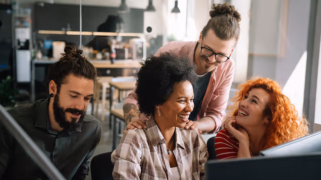 Four diverse coworkers smiling and collaborating around a computer in a modern office.