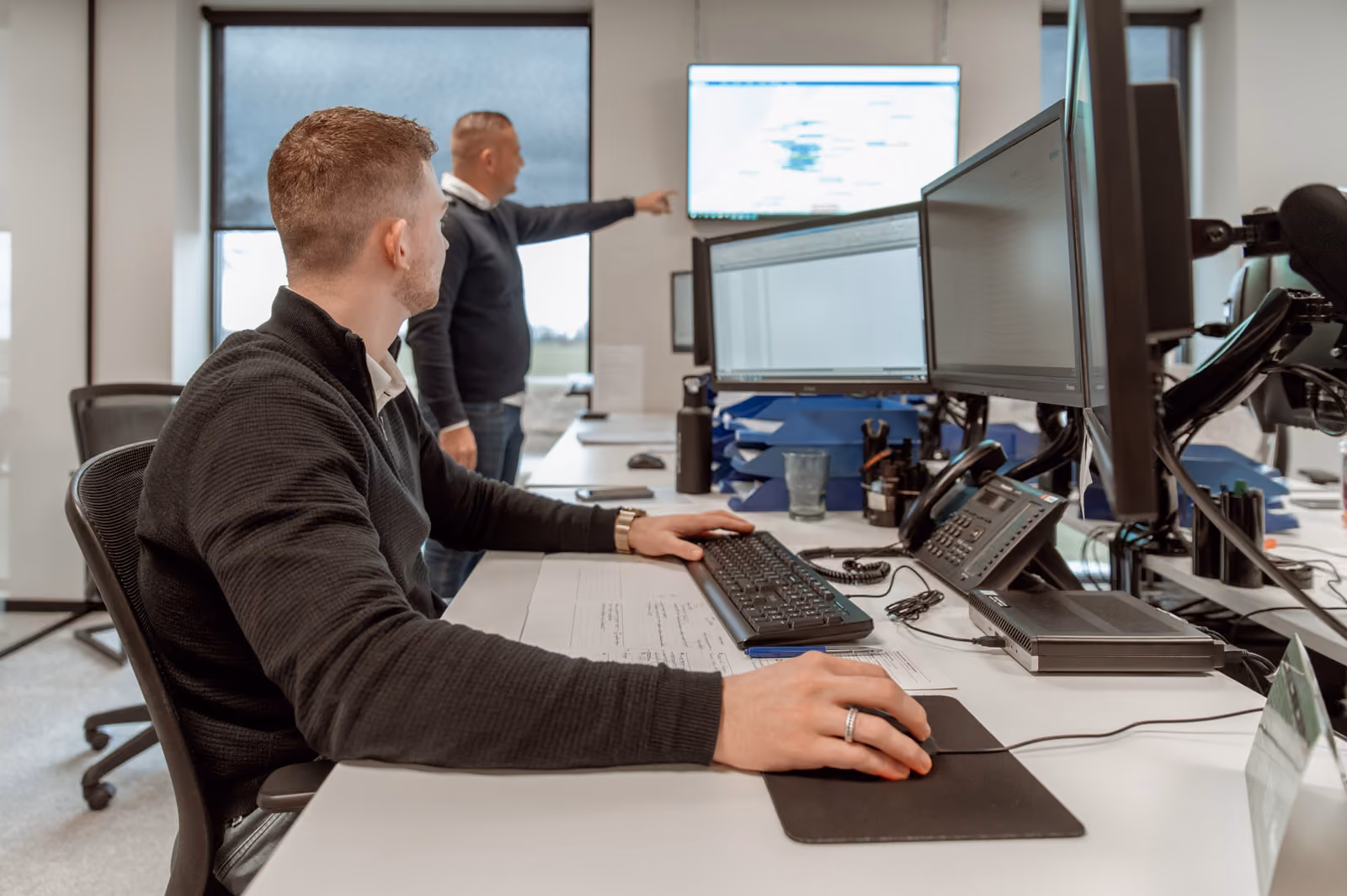 Een man achter de computer kijkt naar een collega's die staat te wijzen naar een scherm wat op de muur hangt bij Solid Systems. 