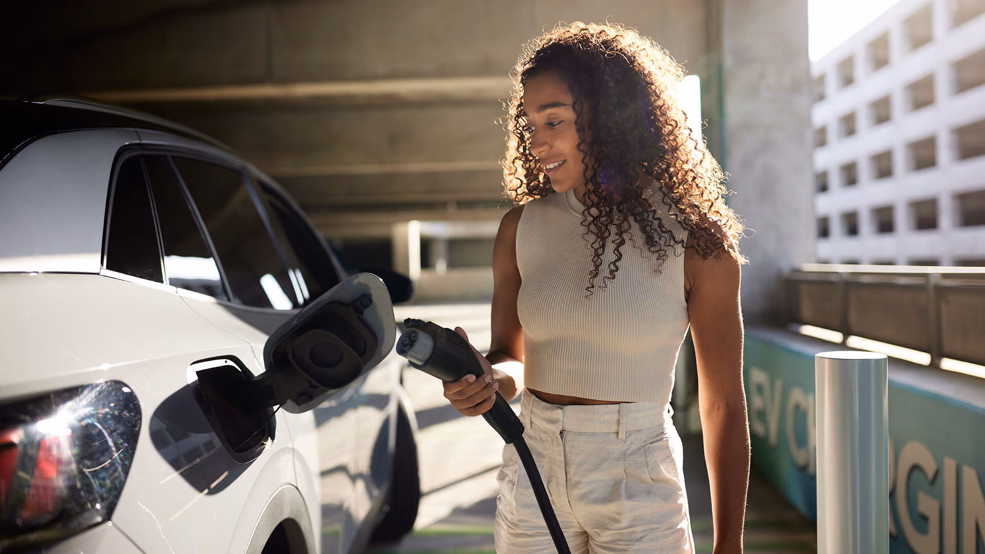 Een vrolijke vrouw met een opvallende bos krulhaar en in het wit gekleeds steekt de laadkabel in haar witte elektrische auto.