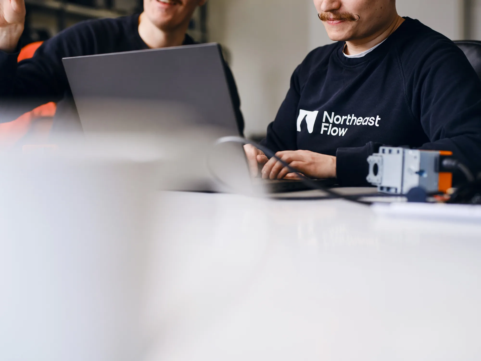 Two men in black shirts working on laptops, one wearing a sweatshirt with the Northeast Flow logo.