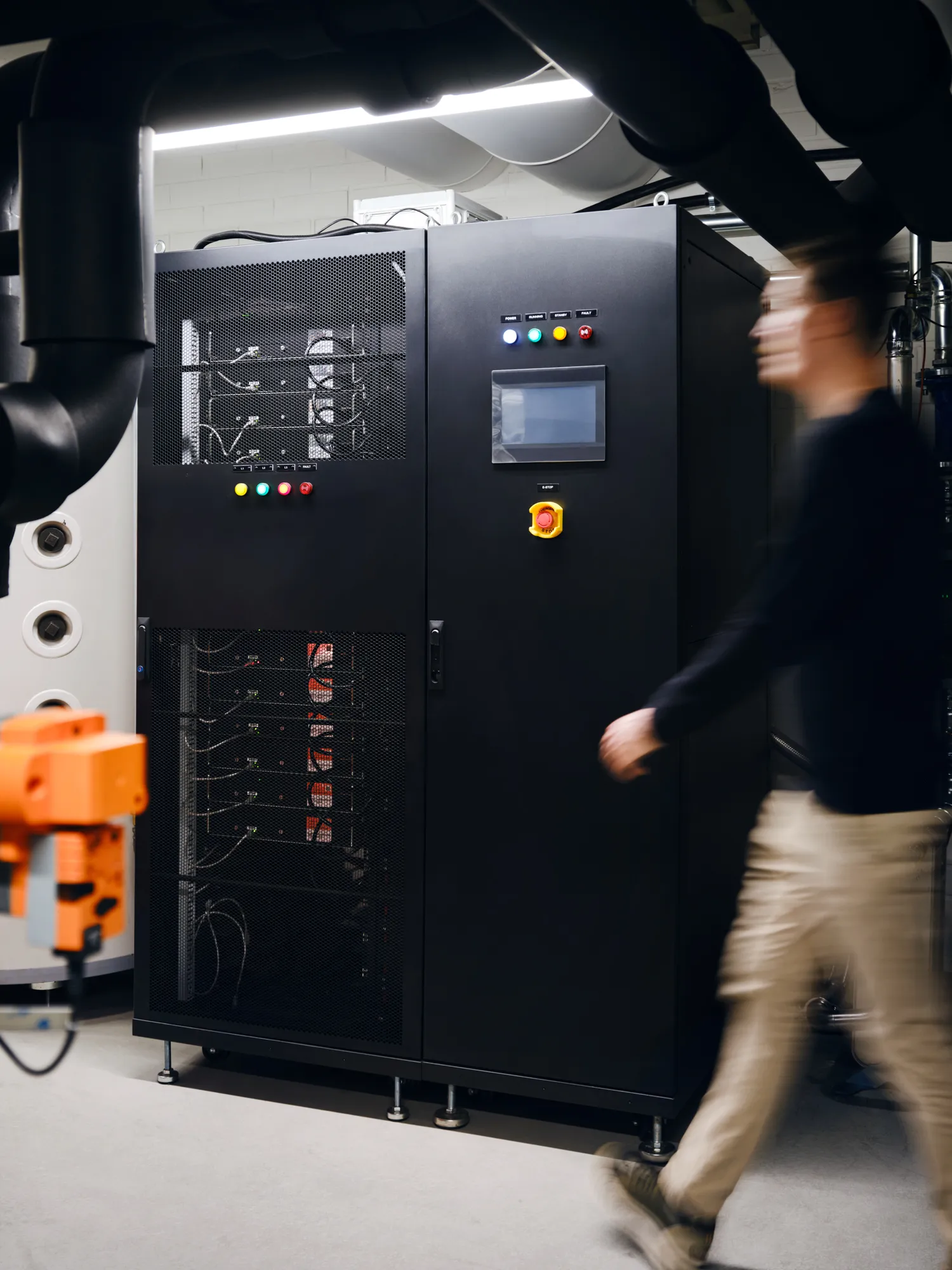 Blurred person walking past a large black industrial control panel with colored buttons and a screen in a utility room.