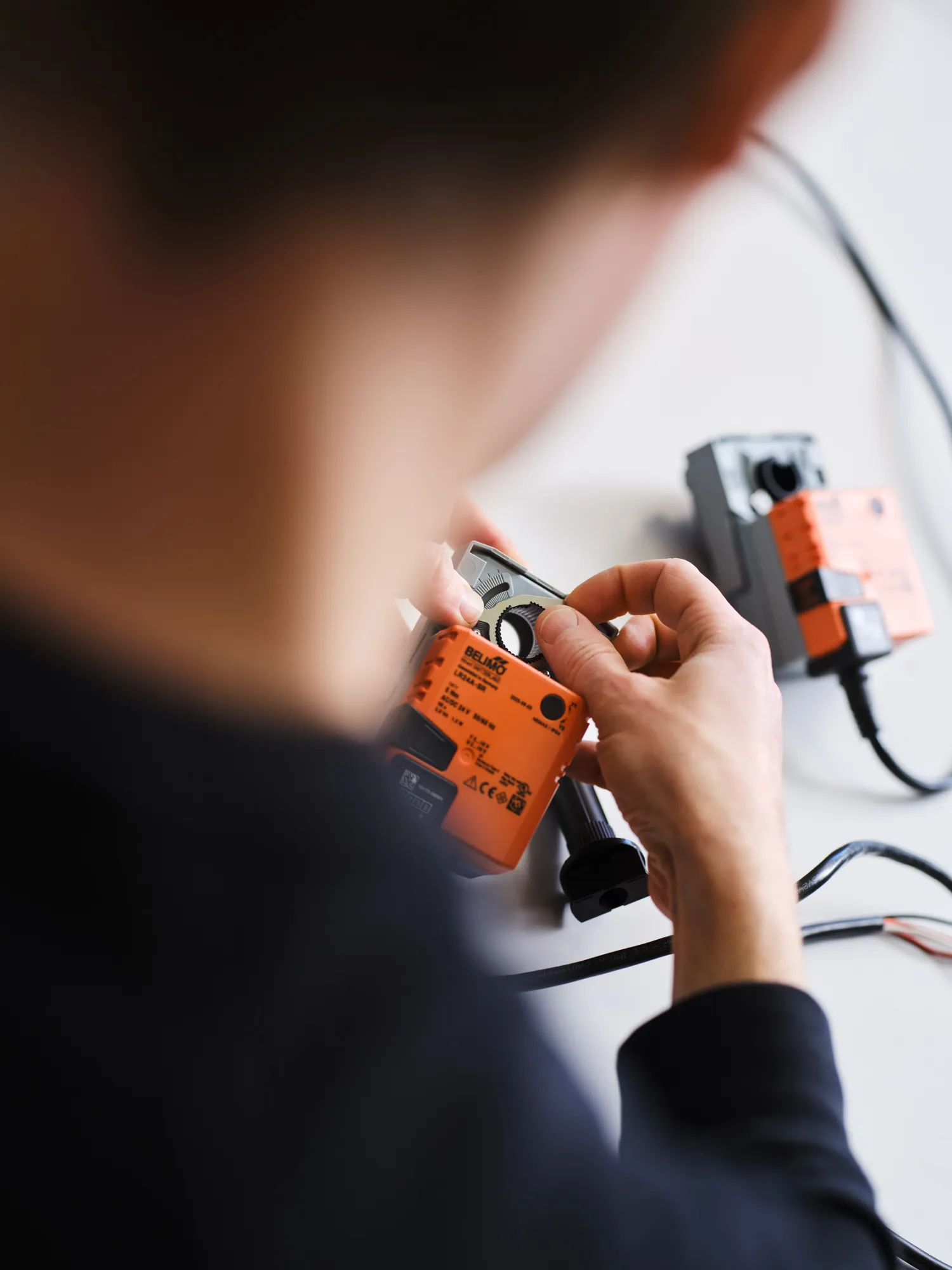 Person adjusting a gear on an orange Belimo electrical actuator with another actuator in the background.