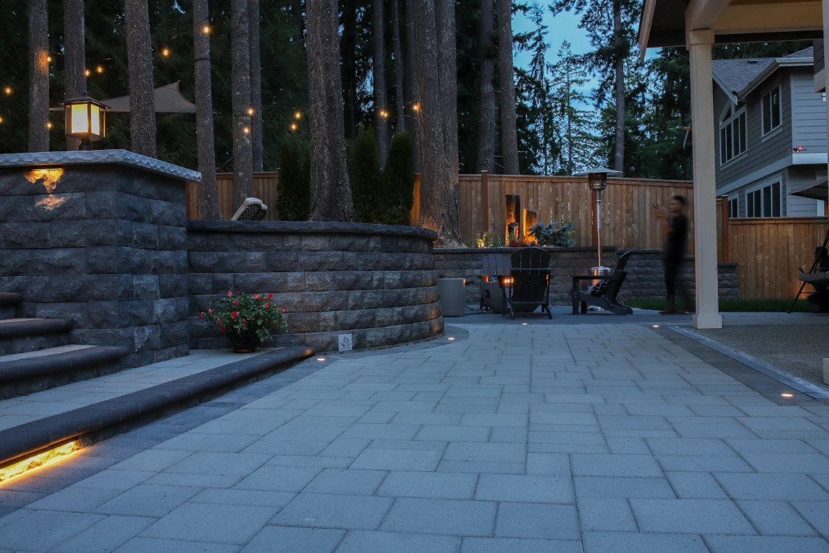 Luxury paver patio with curved stone seating wall, step lighting, and string lights at dusk, outdoor living design by Angkorscape in Greater Seattle