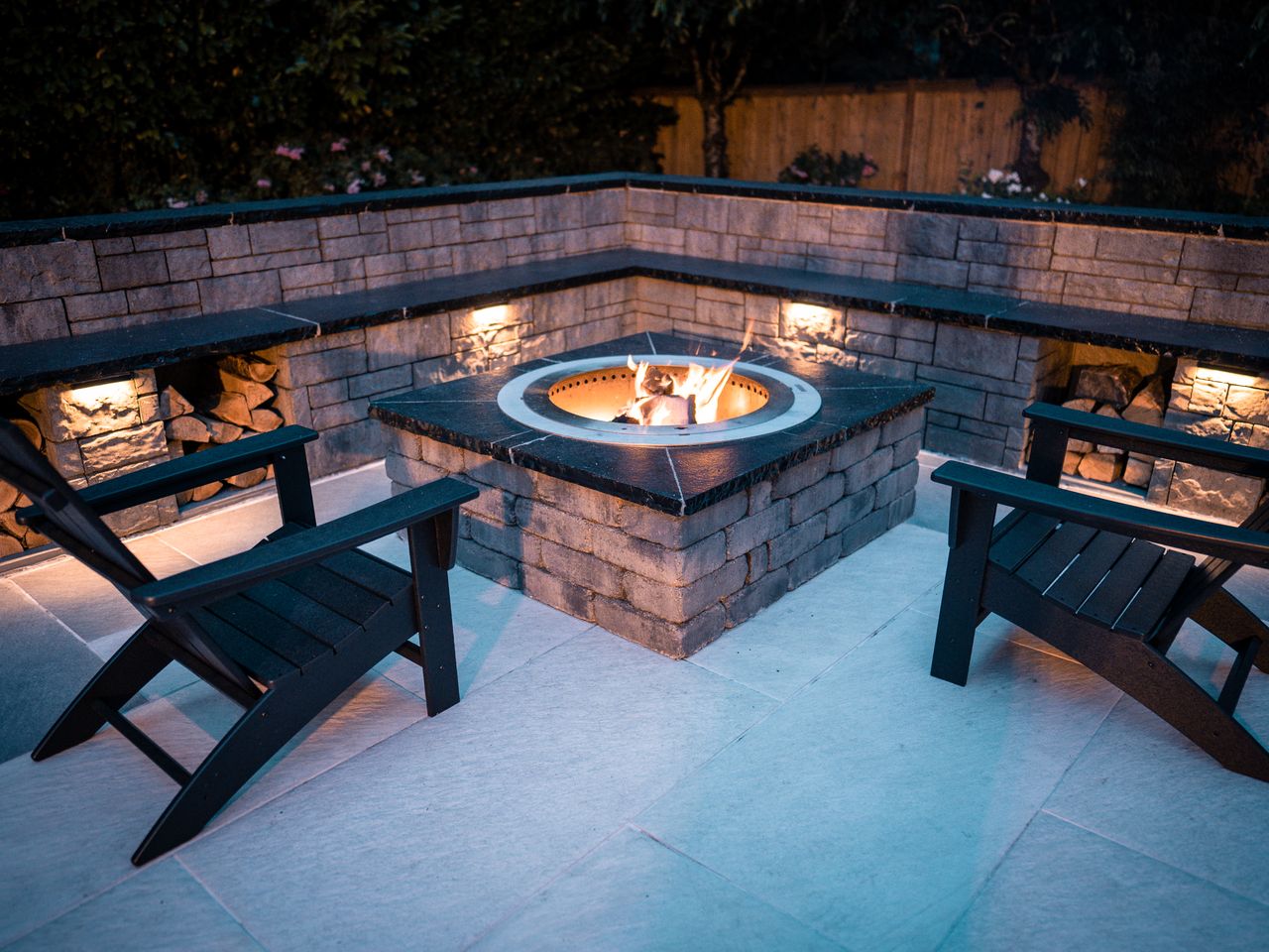 Fire pit flanked by two wooden chairs near a retaining wall with landscape lighting