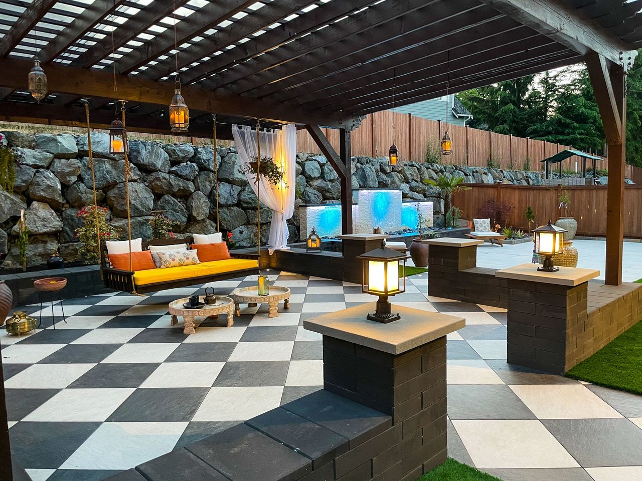 Suspended swing seating beneath a pergola, with a low tray-style table and lit stone pillars