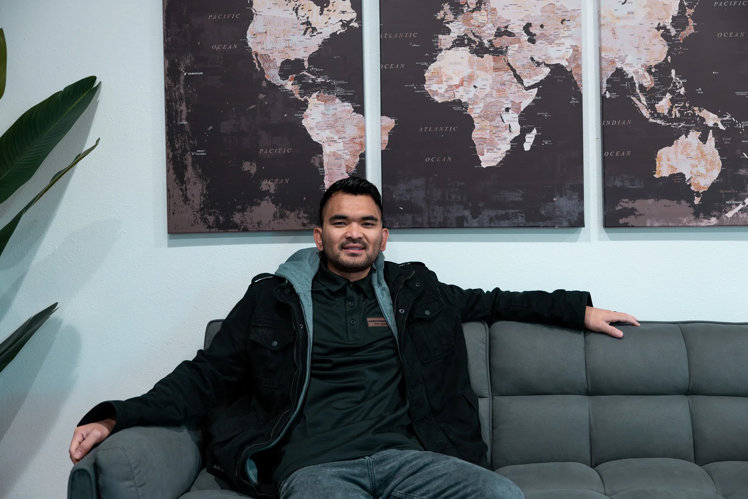 Man in dark jacket sitting on a gray couch with a three-panel world map on the wall behind him.