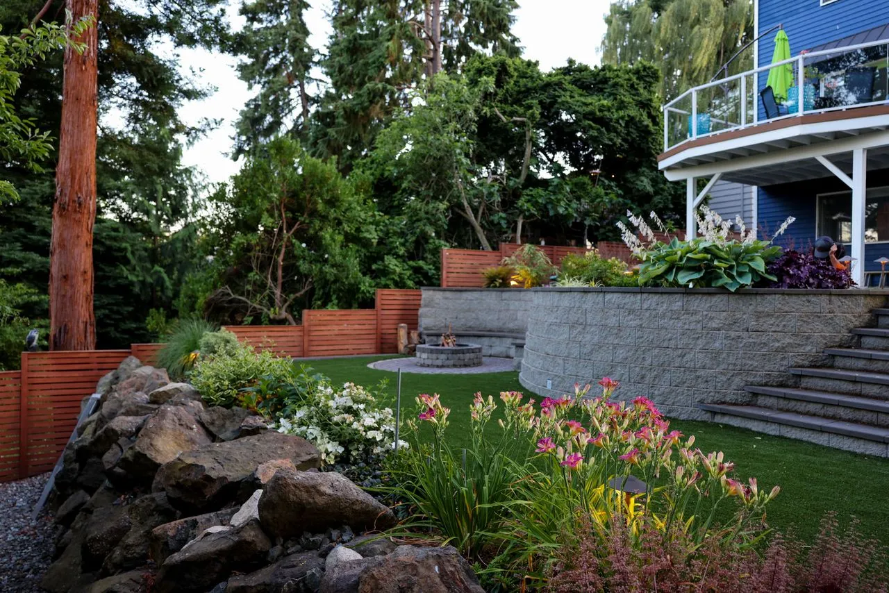 Sloped backyard landscaping with a tiered retaining wall, layered garden beds of plants and flowers, and a fire pit seating area.
