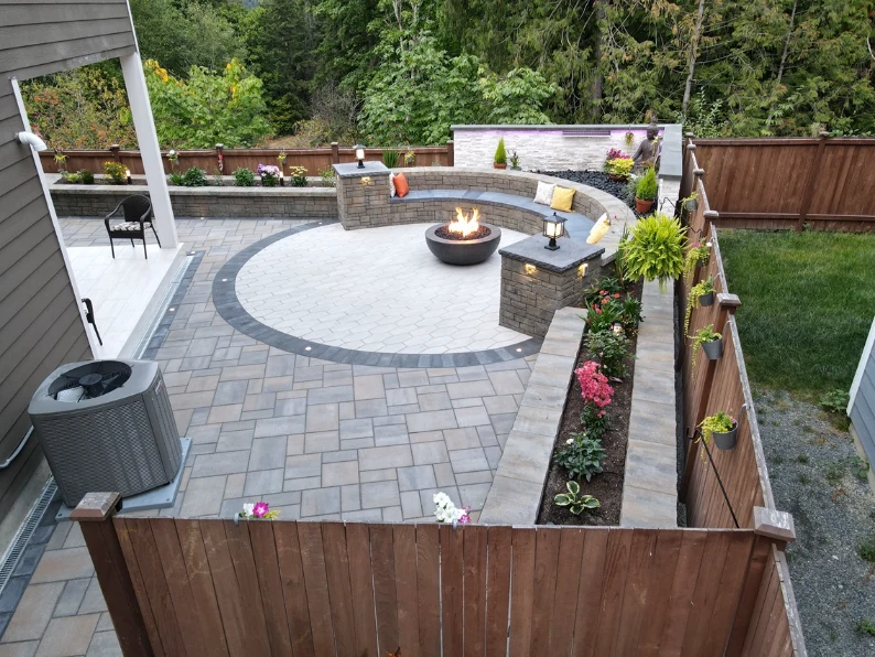 Circular paver patio with a round fire bowl, curved stone seat wall, lit pillars, and a raised flower planter