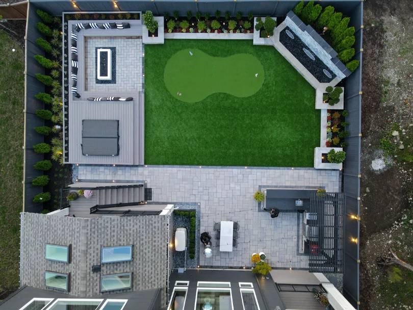 Aerial view of a paver patio, composite deck with fire table, putting green, and stone water feature wall