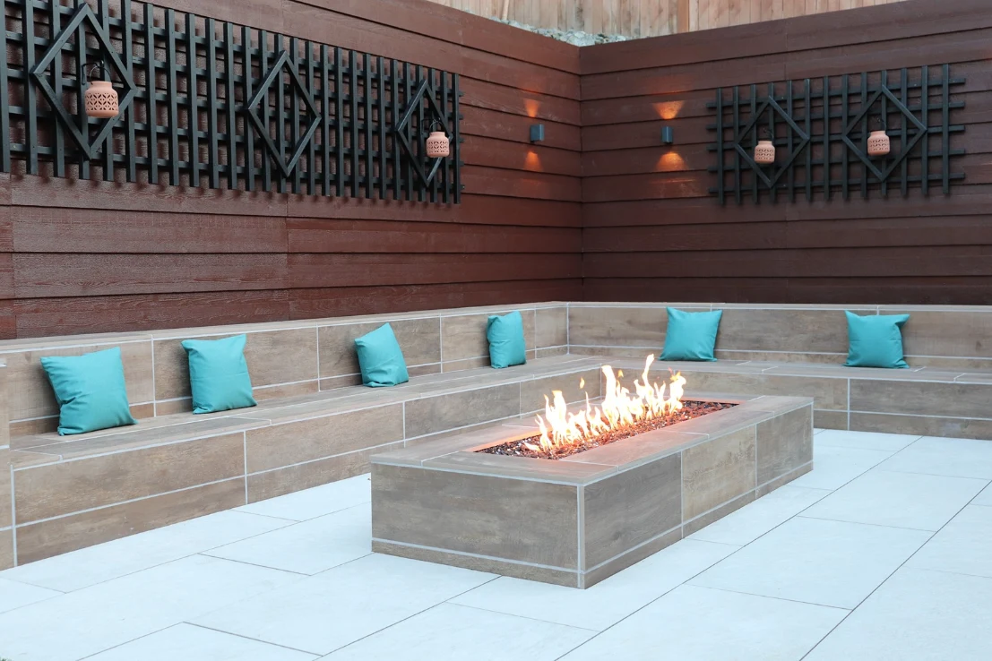 Linear gas fire pit with wood-look tile bench seating, teal cushions, and a cedar privacy fence with black trellis panels