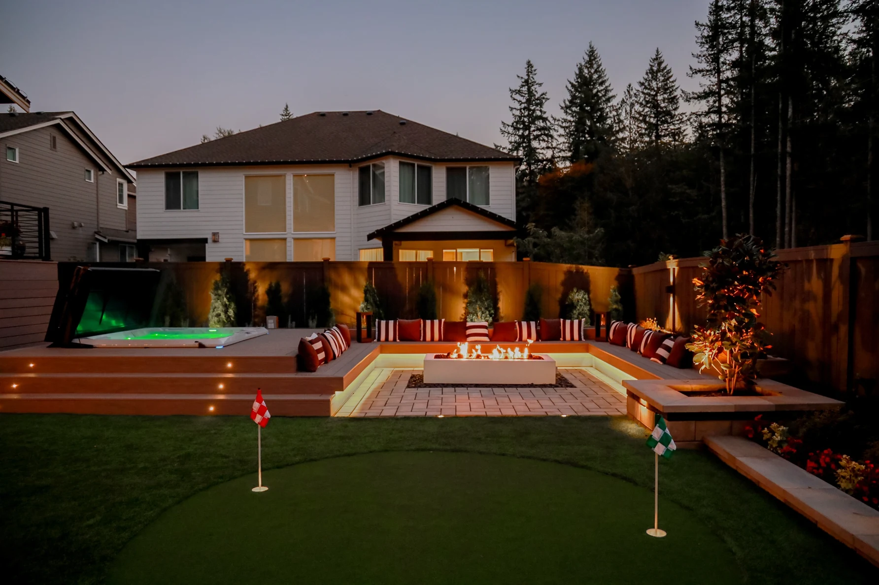 Backyard at dusk with tiered seat walls, sunken fire pit lounge, hot tub deck, and a lit putting green