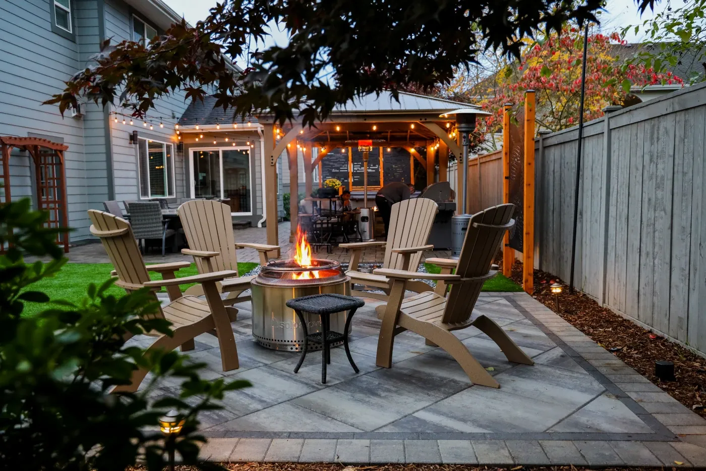Evening photo of a completed paver patio with Adirondack chairs around a fire pit and a lit wood pergola in the background