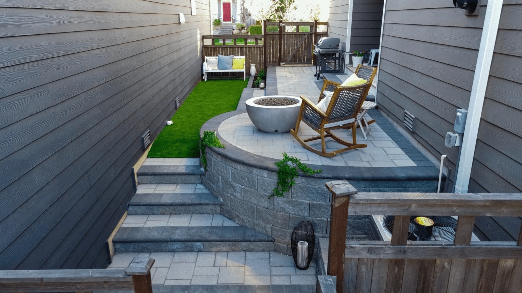 A compact urban backyard transformed with tiered stone paver steps, a circular fire pit seating area with rocking chairs, a strip of artificial turf, and a wooden privacy gate, showcasing a modern minimalist outdoor living design.