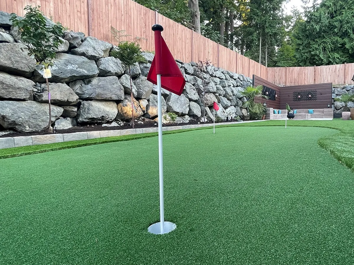 A backyard putting green with red golf flags, a natural boulder retaining wall with planted shrubs, and a wood privacy fence surrounded by tall evergreen trees.