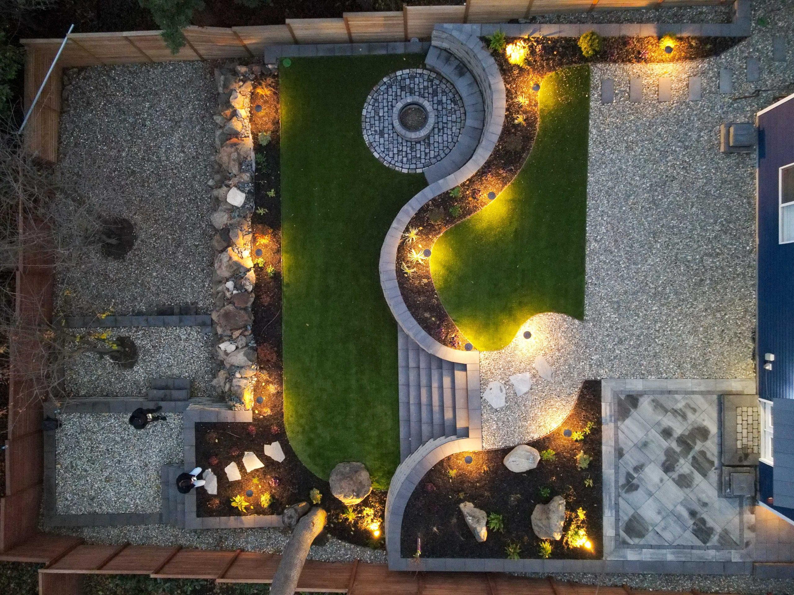 A professionally landscaped backyard at dusk featuring a circular fire pit seating area with Adirondack chairs, curved stone retaining walls, string lights strung between tall evergreen trees, a hammock area, and warm pathway lighting throughout.