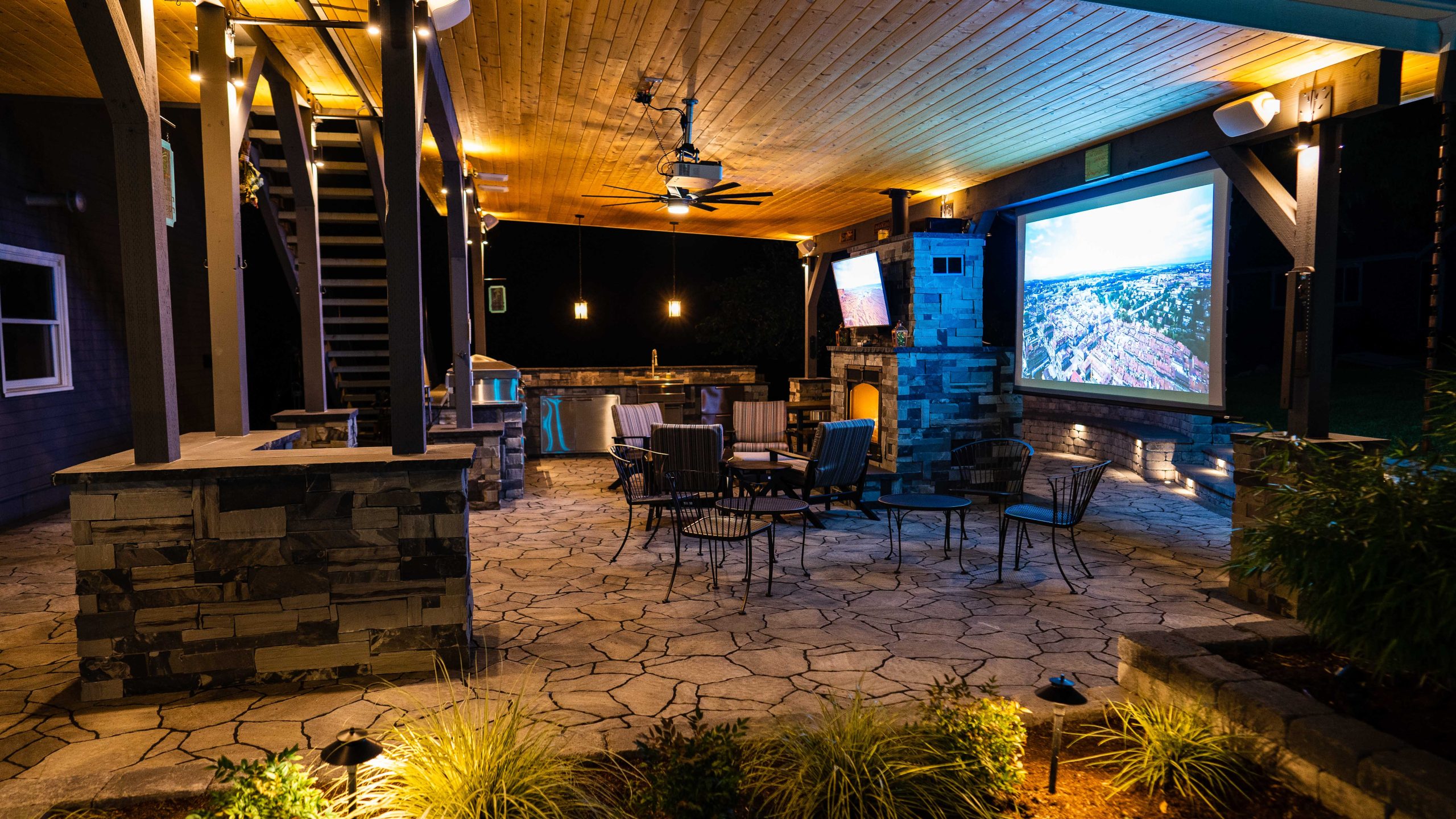 A covered outdoor living space at night featuring a stone fireplace, large projection screen, mounted TV, outdoor kitchen with bar seating, string lights, wooden ceiling with ceiling fan, and stone tile flooring.