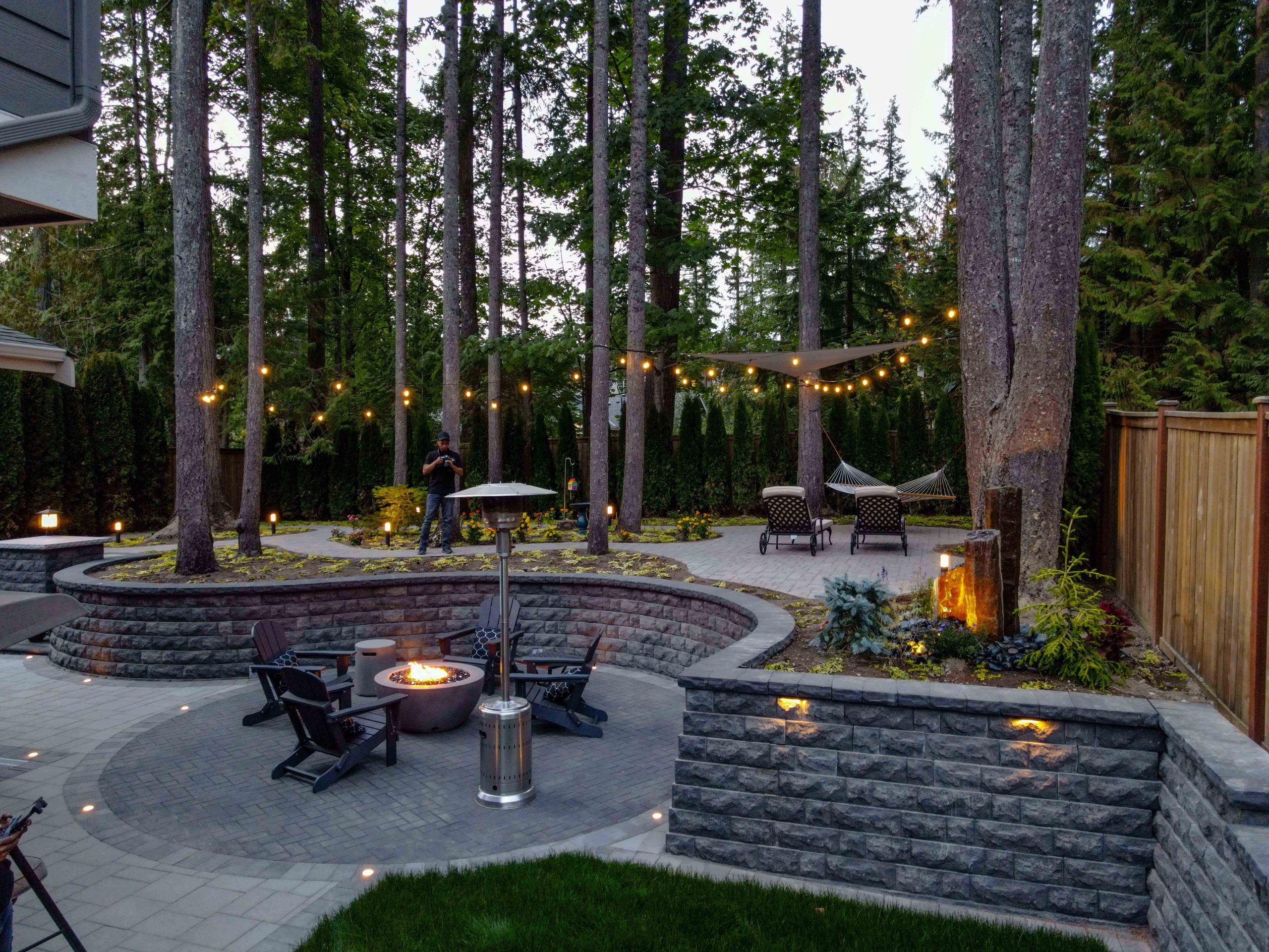 An aerial night view of a custom landscaped backyard featuring a sinuous artificial turf lawn with a circular cobblestone fire pit, curved illuminated retaining walls, gravel pathways, decorative boulders, and warm landscape lighting throughout.