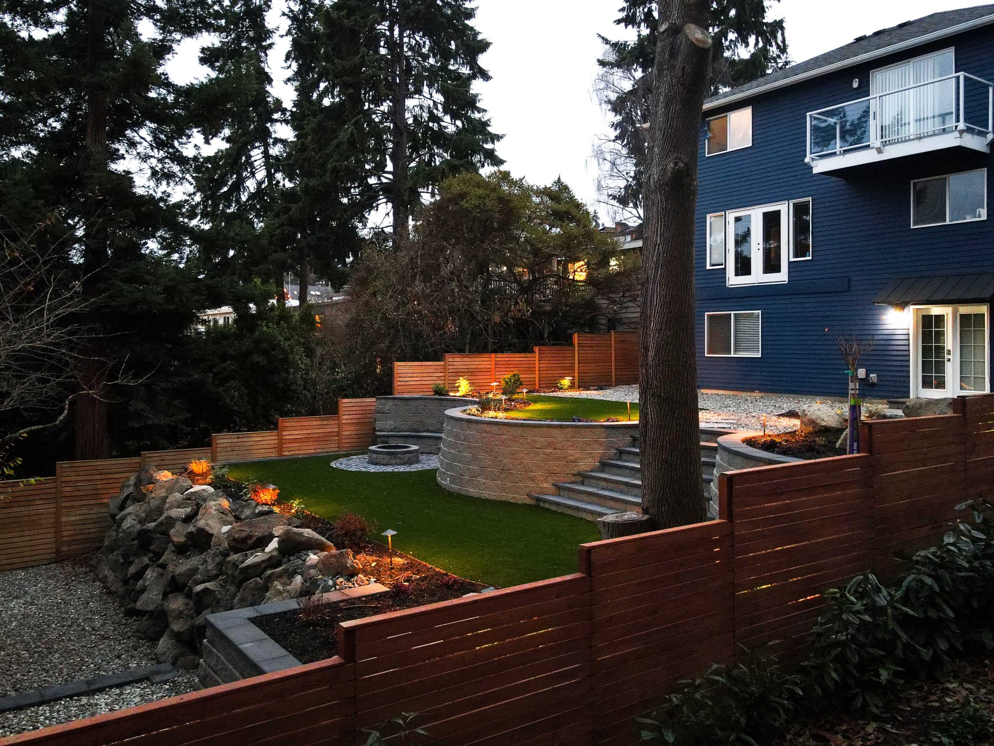 A modern backyard outdoor living space at night featuring a green-lit hot tub on a composite deck, a sunken fire pit lounge area with built-in bench seating and striped cushions, warm LED under-bench lighting, and wall-mounted sconces illuminating a wooden