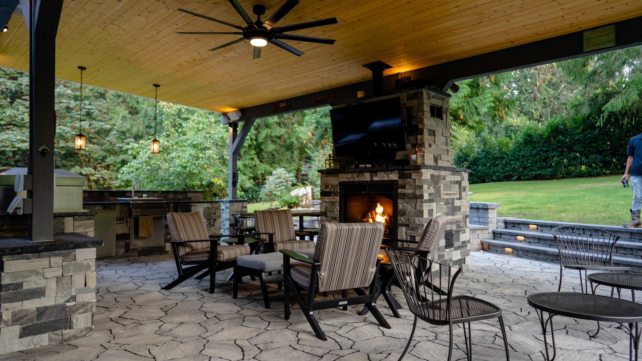 A luxury covered outdoor living space featuring a stone fireplace with a mounted TV, striped cushioned seating, an outdoor kitchen with stainless steel appliances, pendant lantern lighting, a large ceiling fan, and a view of a lush green backyard lawn.