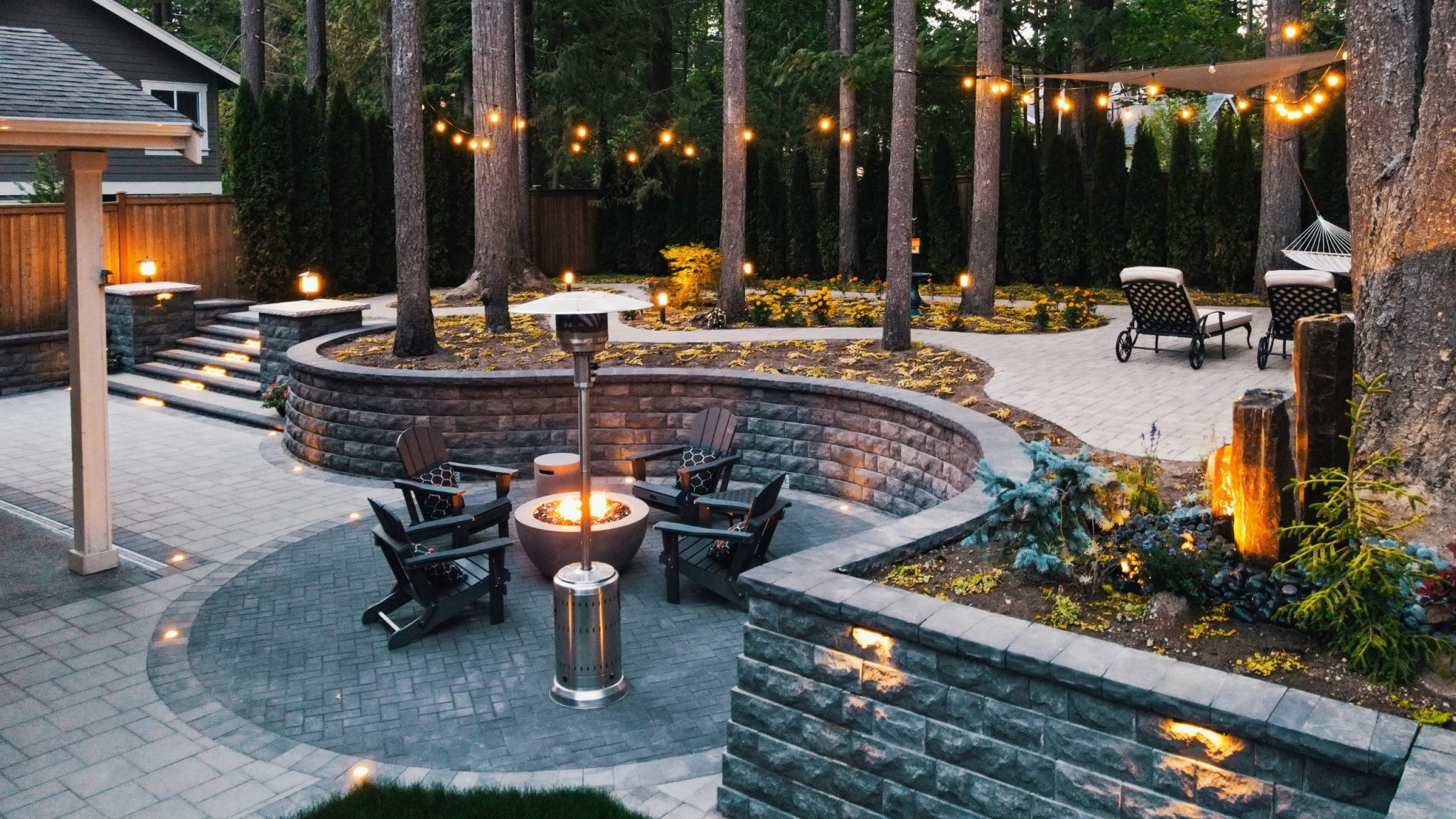 A luxury backyard patio at dusk featuring a circular fire pit seating area with Adirondack chairs, curved tiered stone retaining walls with step lighting, string lights strung between tall evergreen trees, a hammock, lounge chairs, and illuminated ornament