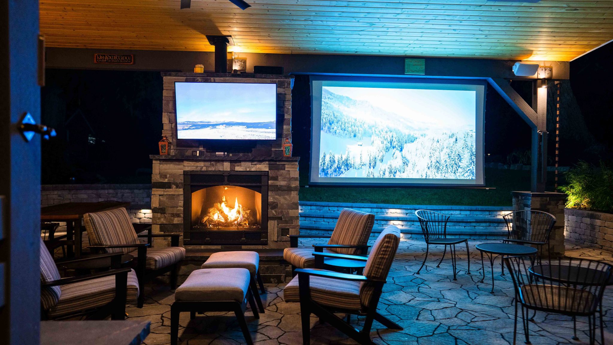 A covered outdoor living space at night featuring a lit stone fireplace with a mounted TV, a large projection screen displaying a snowy mountain landscape, striped cushioned lounge chairs with ottomans, stone tile flooring, and warm ambient ceiling lightin