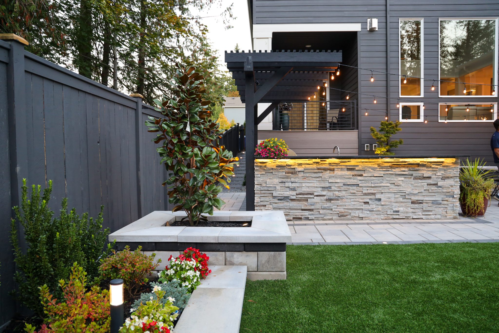 A professionally landscaped backyard at dusk featuring a raised garden bed with a magnolia tree and red and white flowers, an illuminated stacked stone outdoor bar counter, a dark pergola with string lights, a lush artificial turf lawn, and pathway bollard