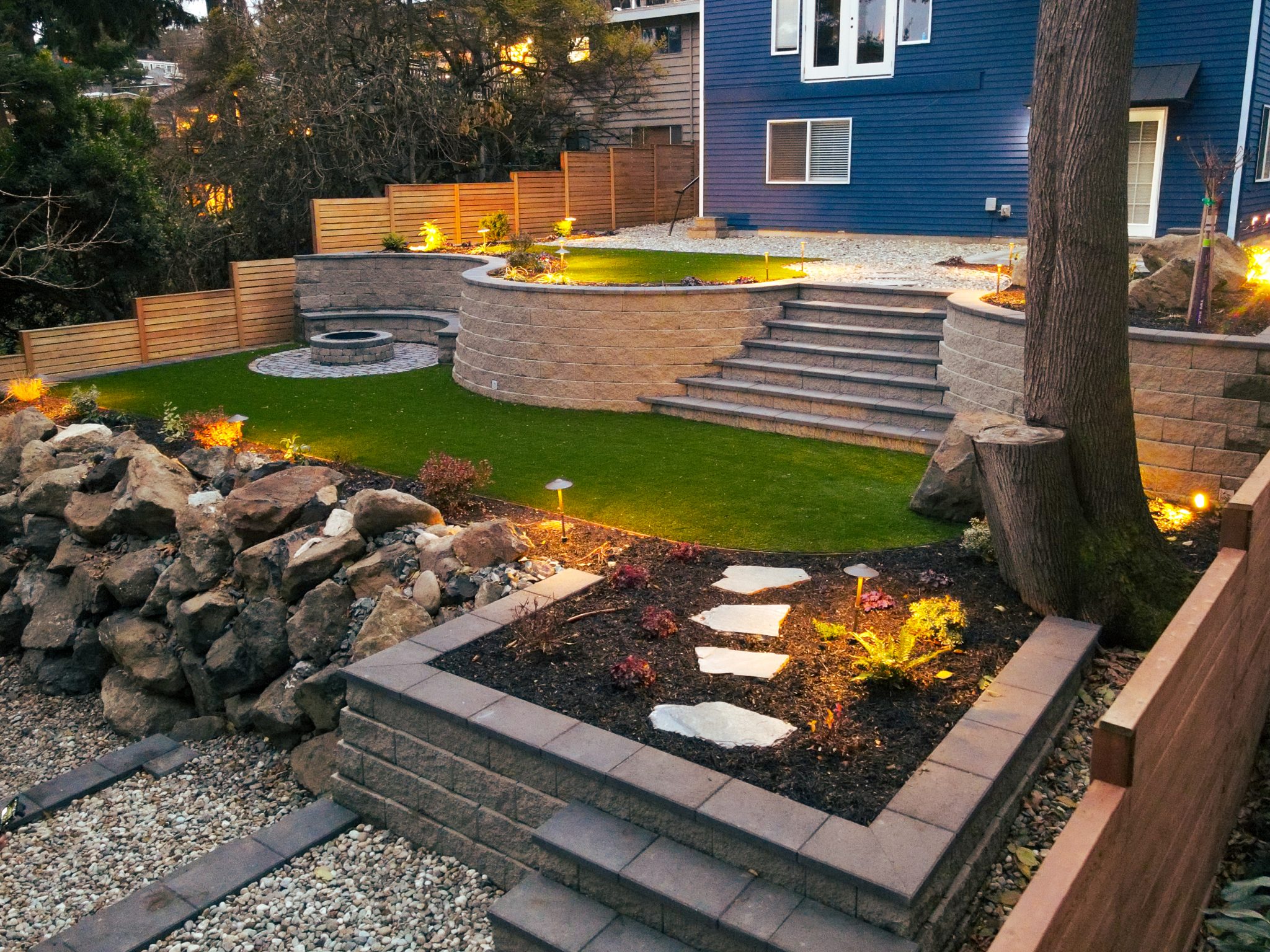 A professionally landscaped backyard at dusk featuring tiered stone retaining walls, a circular fire pit on artificial turf, illuminated garden beds with pathway stake lights, natural boulder accents, stepping stone walkways, and a horizontal slat wood pri
