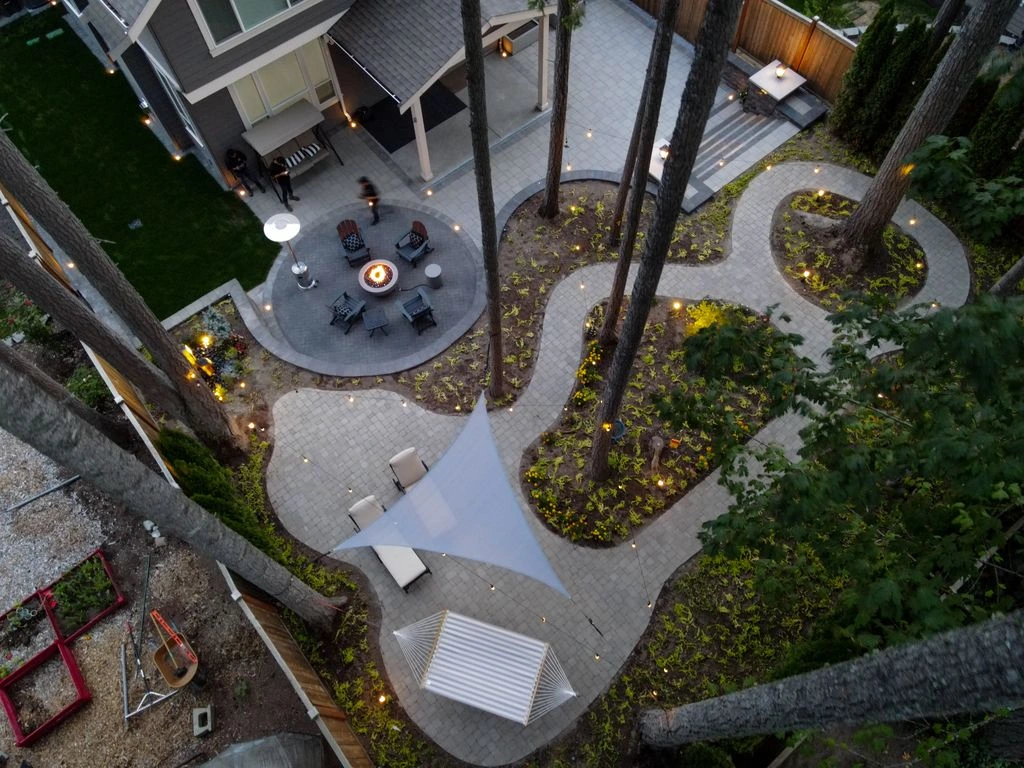 Aerial view of a Snoqualmie backyard with a curved stone patio, fire pit, shade sail, and pine trees.