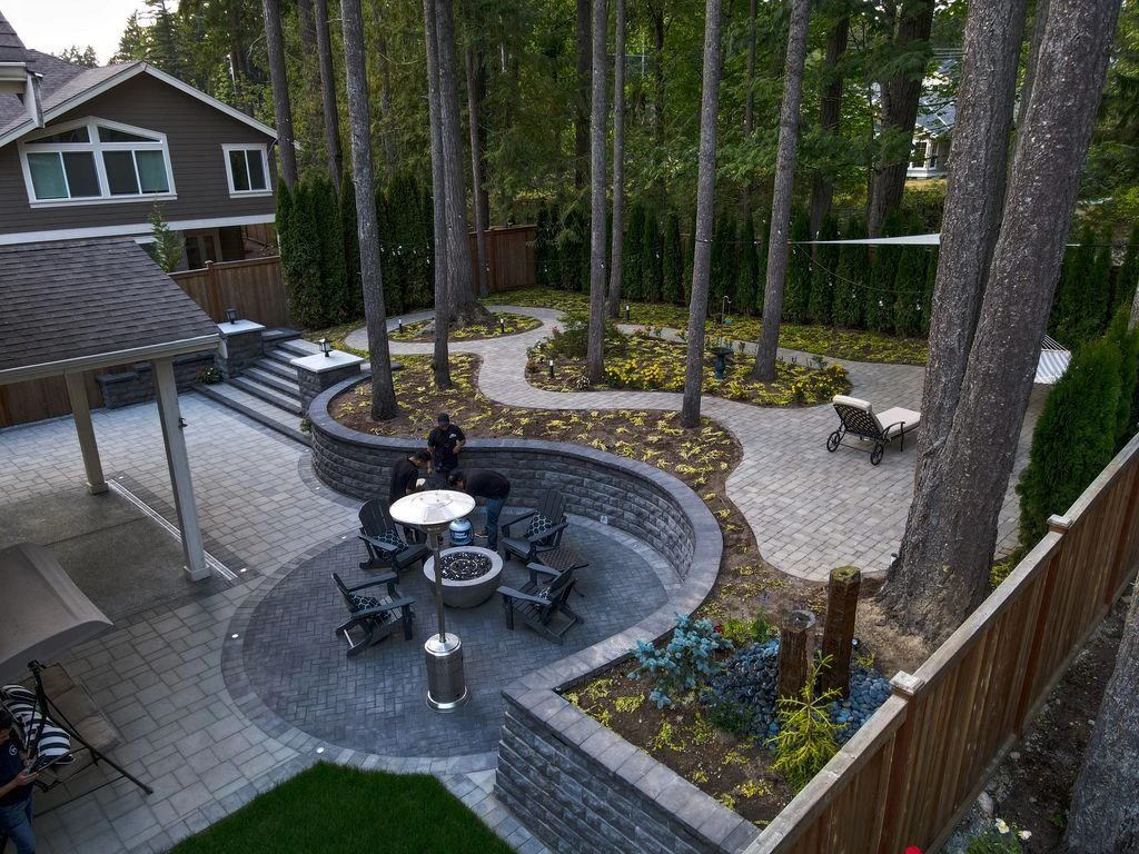A completed Snoqualmie project with a stone patio, fire pit, retaining wall, and garden beds among pine trees.