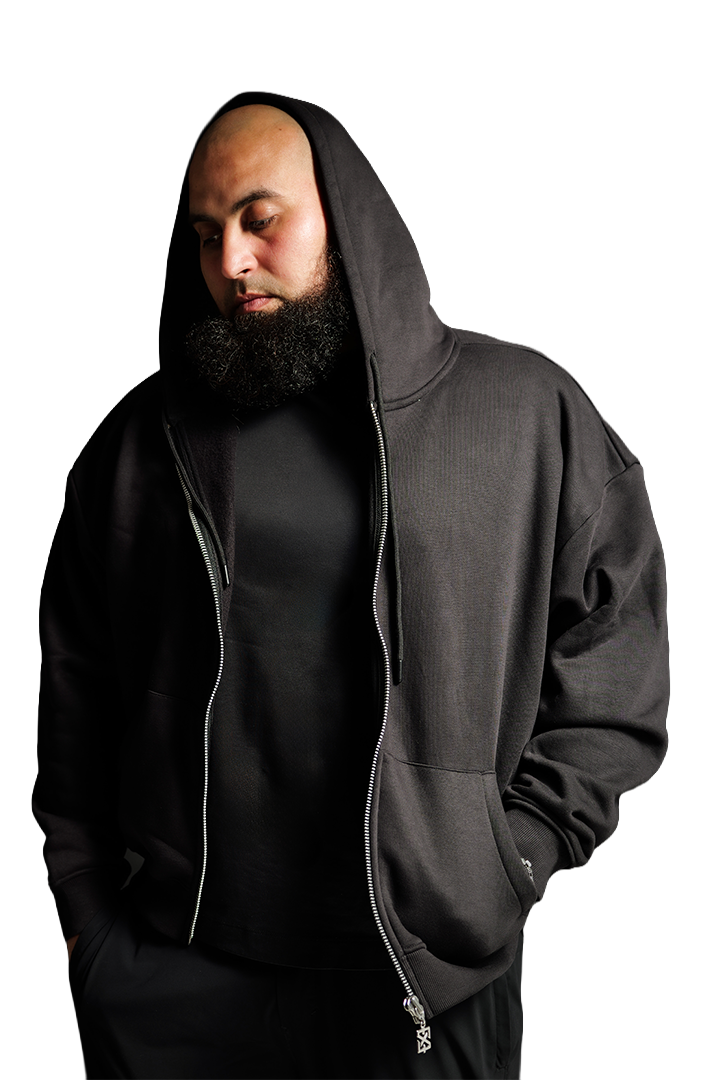Man met een zwarte hoodie met capuchon en een volle baard, kijkt naar beneden tegen een zwarte achtergrond.