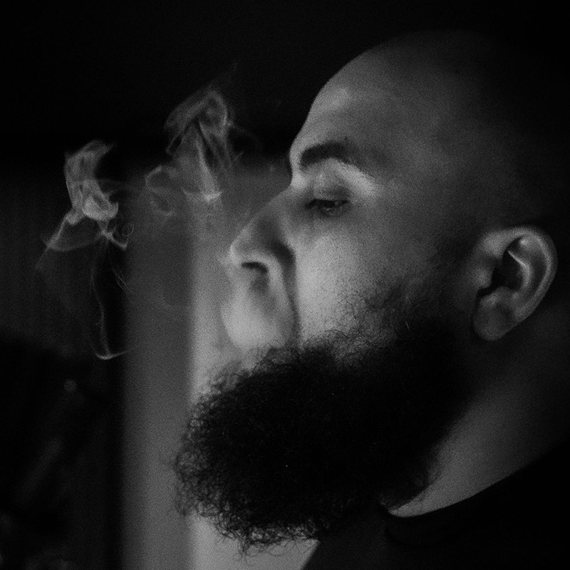 Black and white close-up portrait of AfroTura with eyes closed, exhaling smoke against a dark background