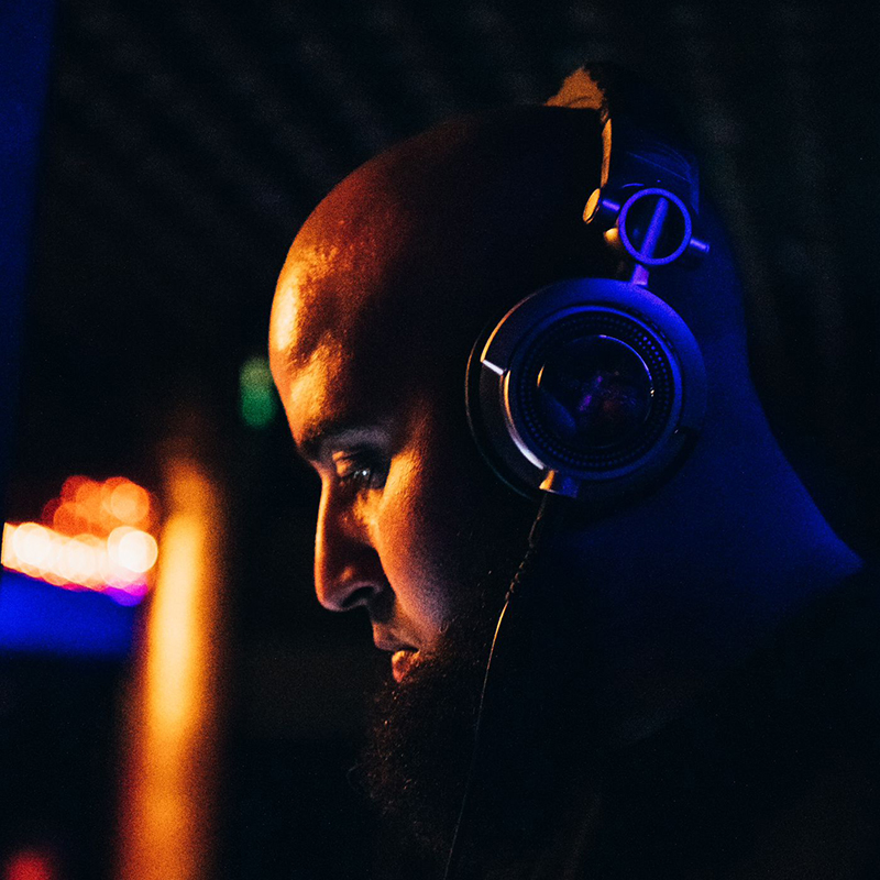 Side profile portrait of AfroTura wearing headphones, lit by deep blue and orange light during a live DJ performance